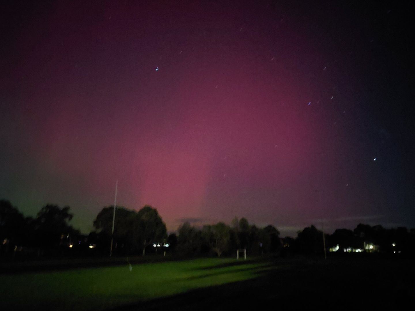 Another photograph taken at night showing an aurora above dark trees and fewer lights. The aurora is an orangey-red in the centre, with the brightness reducing out towards the edges of the frame. The football field where the photo was taken is gently illuminated from a building behind the camera.
