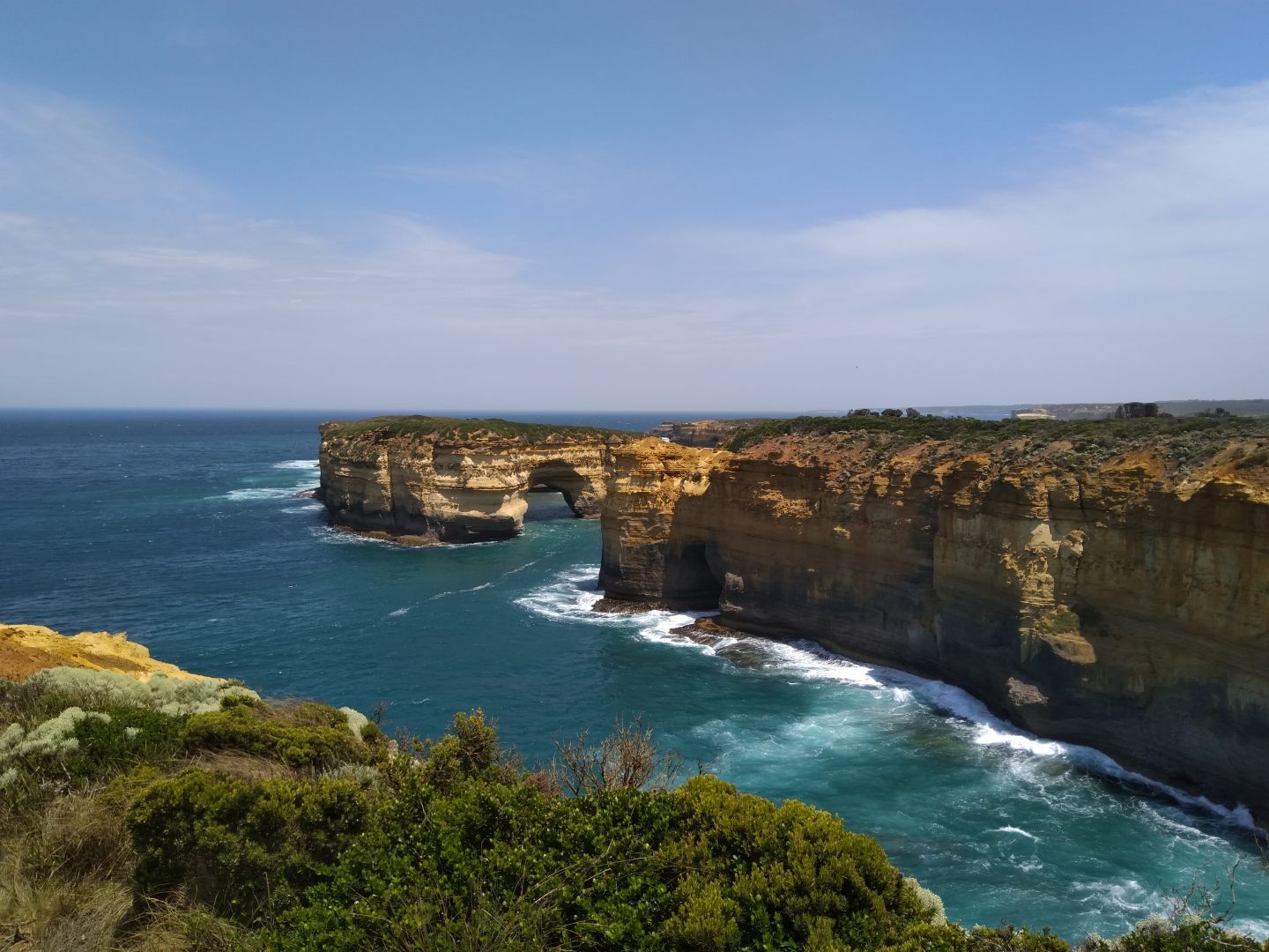 A rocky golden peninsula extends out into the vibrant blue sea. At its end, like the tittle of an 'i', lies an island with a green bushy carpet on top, and an arch over the blue sea beneath