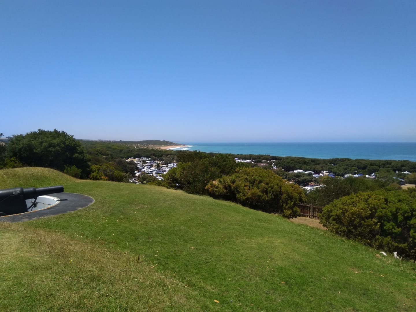 A large early 20th-century gun sits behind grassy fortifications. Below the hill, houses sit near the edge of the sea, surrounded by green trees