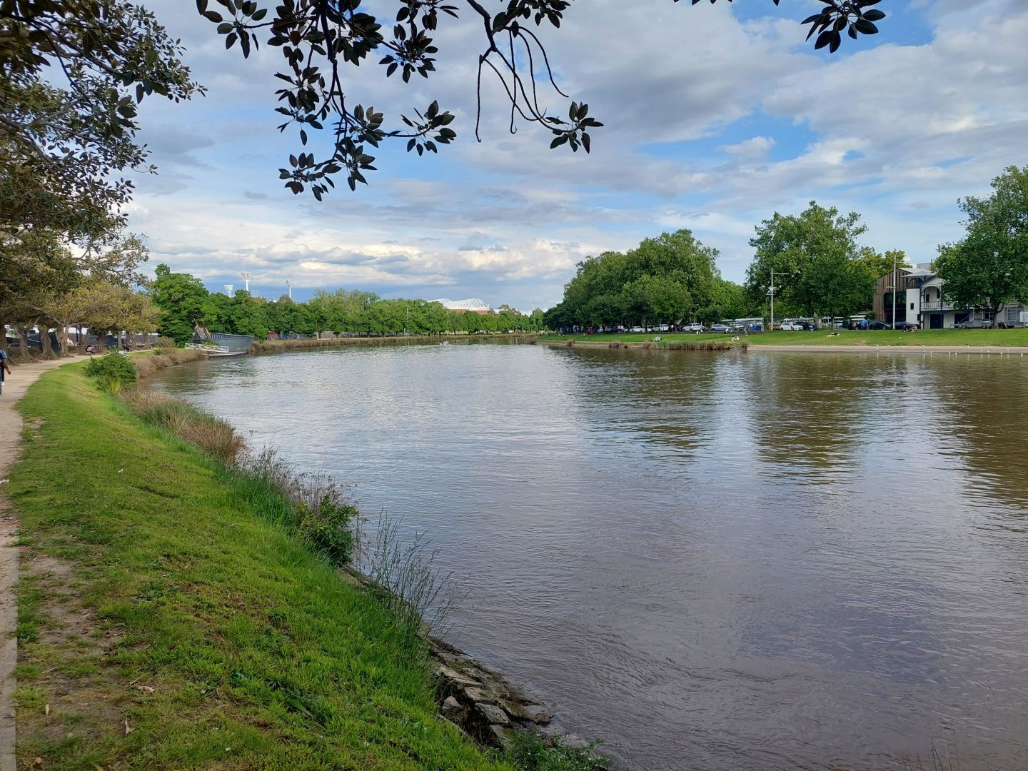 Looking out over the Yarra River, which runs through the heart of Melbourne out to the east. In this section, it bows out as it curves between the sporting precincts (Melbourne, Olympic, and Yarra Parks) and the Royal Botanical Gardens and associated parklands. The river itself is slightly brown, but doesn't look too bad on this particular day. The bank on which the picture was taken has a tan gravel walking track along the edge of the river as far as the eye can see. In the background, the massive light towers of the Melbourne Cricket Ground and the arches supporting the roof of Rod Laver arena can both be seen behind the trees. On the other side of the river, the end of the line of boat sheds can be seen, belonging to the old rowing clubs in Melbourne.