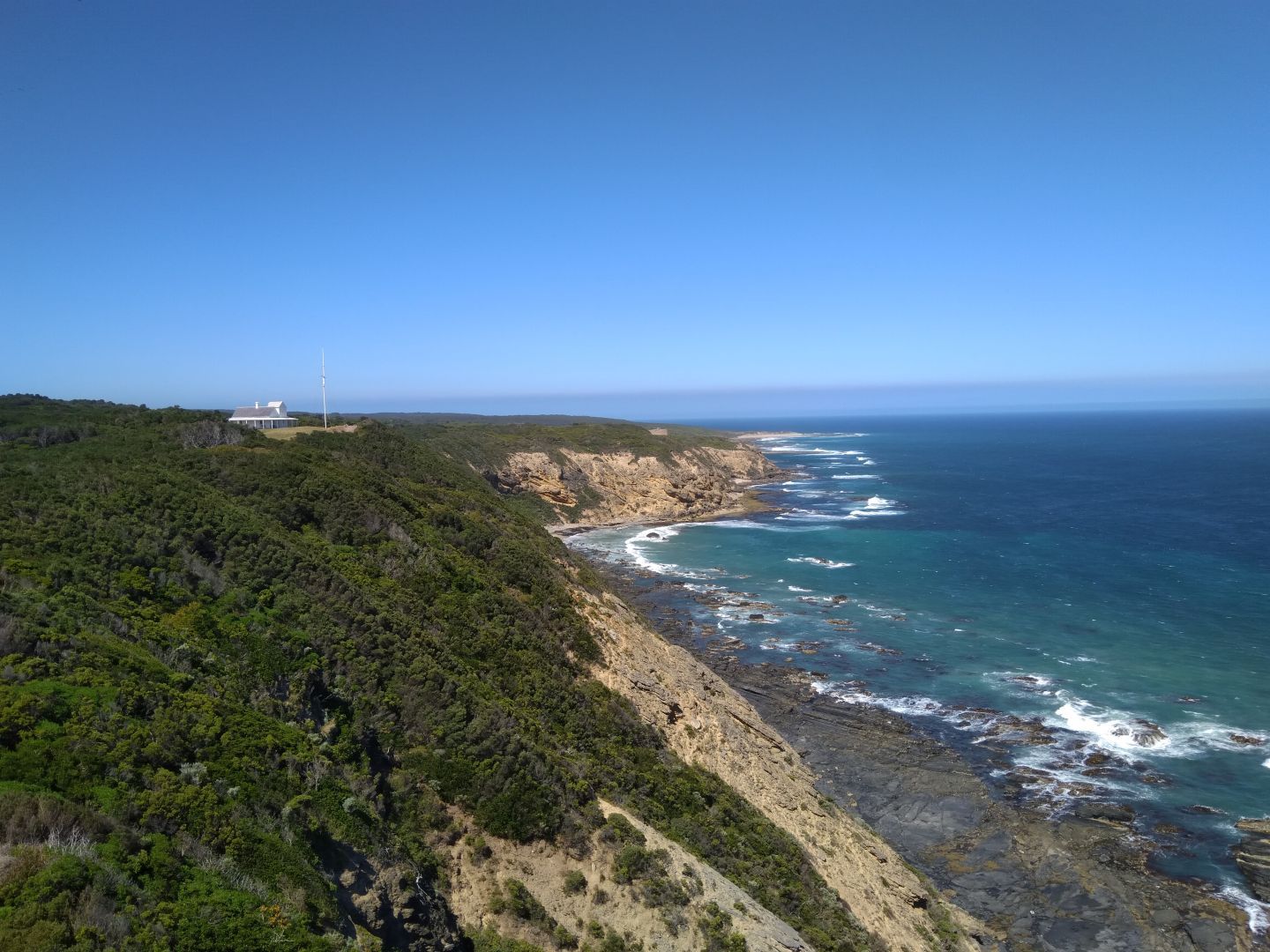 A small house sits on the steep, rocky coast (left) blanketed with thick vegetation. The waves of the deep ocean (right) break on the rocks at the edge of the cliff