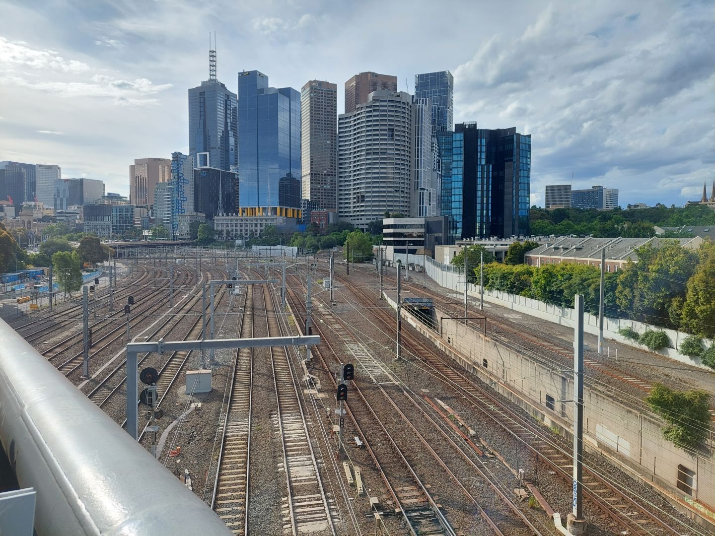 Standing on a bridge over a dozen railway tracks, the photo is facing towards the tall skyscrapers in the Central Business District of Melbourne, Australia. The buildings are monoliths of blue-hued glass, although with some peach and grey concrete towers thrown in for variety. At the front stands the old Herald and Weekly Times Building, a five-story building constructed in 1921 featuring two radio towers on the roof. To the right (east) of the CBD lies a patch of parkland filled with green trees (the Treasury Gardens).