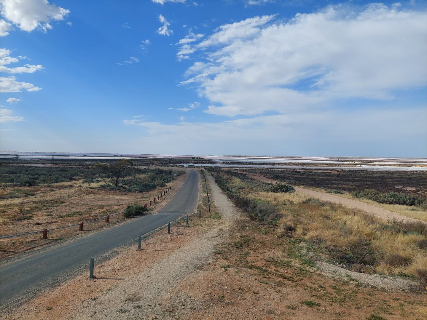 A photo of the lake from a nearby lookout tower. An asphalt road runs from under the lookout towards the edge of the lake. A large proportion of the soil has saltbush growing on it. In the distance, the lake can be seen in pink, blue, and white, under a cloudy blue sky.