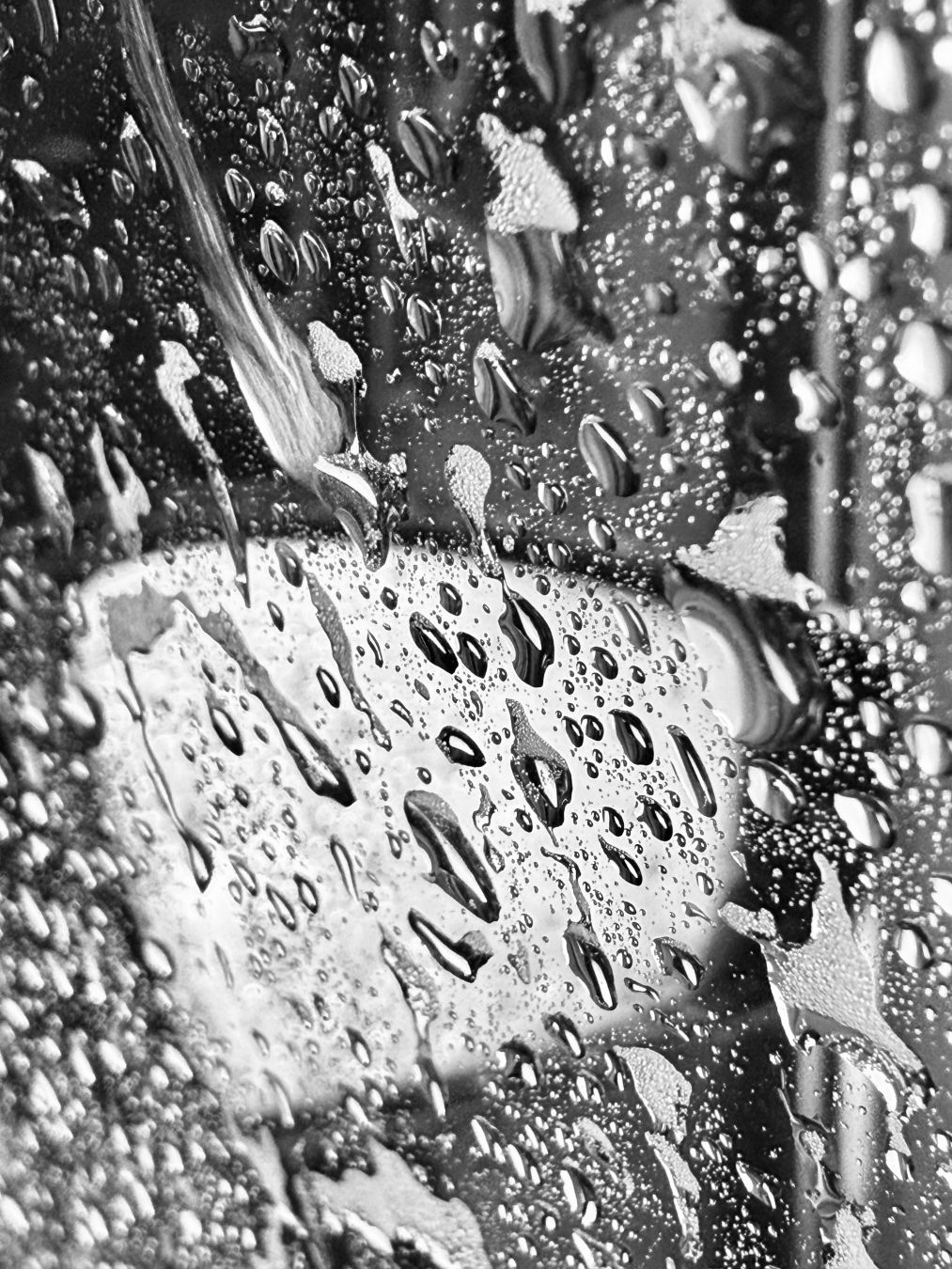 A black and white photograph taken from inside a car at a car wash. Splashes of water and soap streak across the window. A rear view mirror is visible through the water stained window.