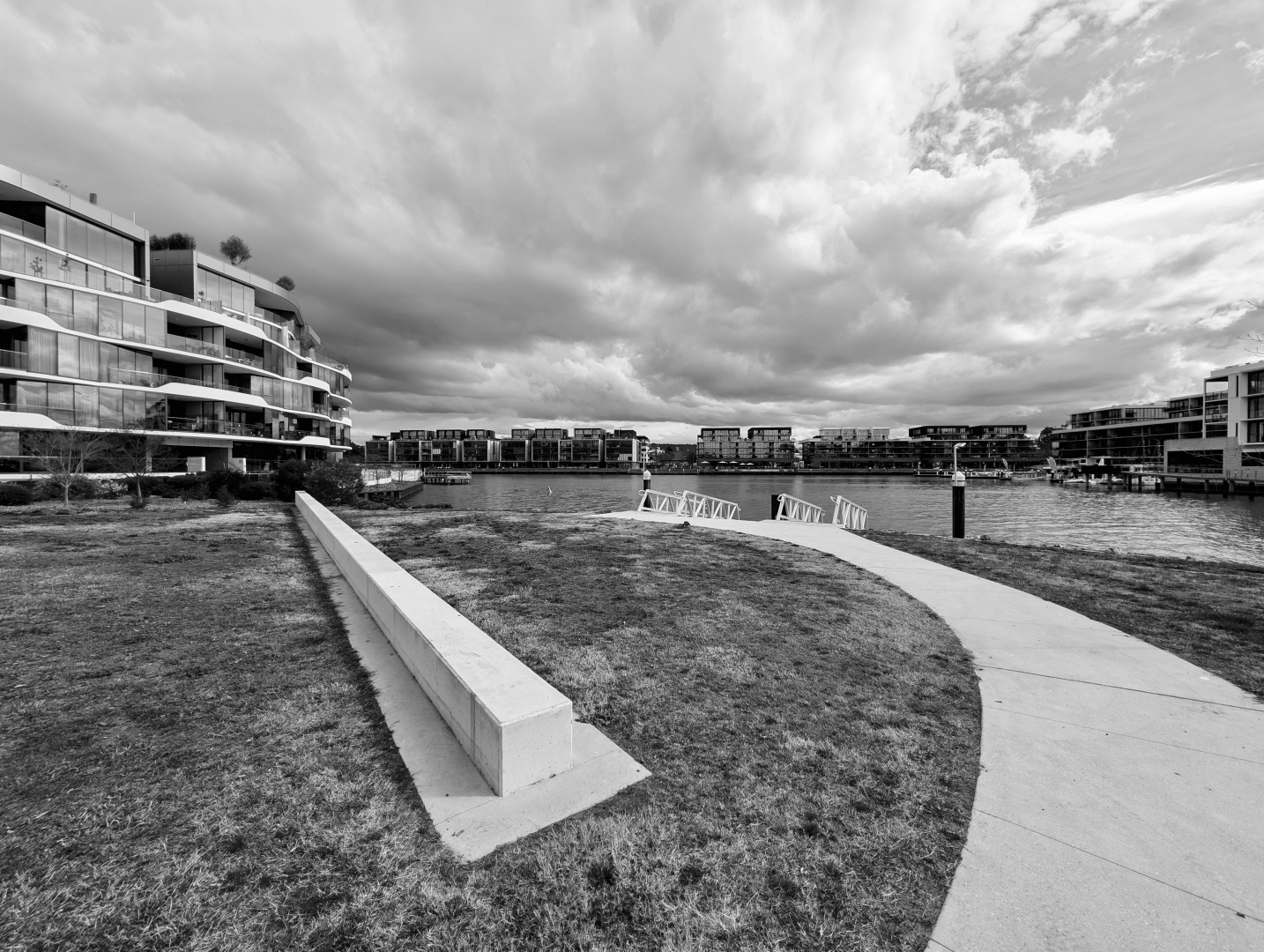 Black and white photograph looking across a park and harbour.  In the foreground is a patch of grass, a concrete path curving into the middle of the frame and a low concrete wall angling left.  On the left and in the distance are blocks of apartments.  Above is an ominous cloudy sky.