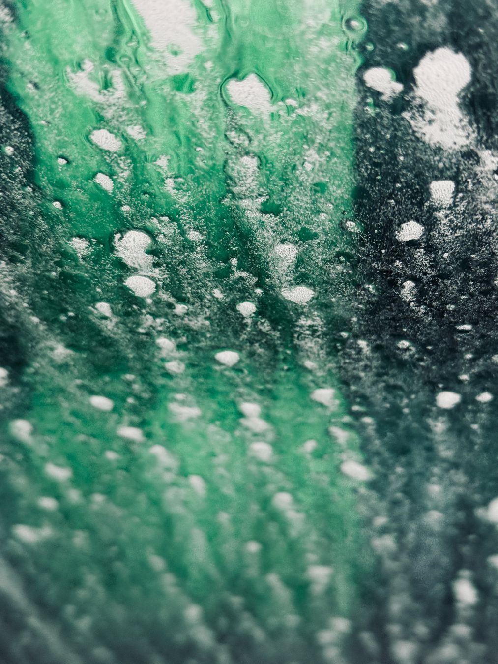 A photograph taken from inside a car at a car wash. Splashes of water and soap streak across the window. Behind is a green blur of brushes spinning.