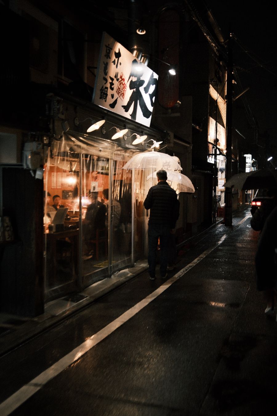 Hungry would be diners roam the wet streets of Kyoto looking for something delicious. They are holding clear plastic umbrellas that gather the light from the restaurant that they are stopped outside of.