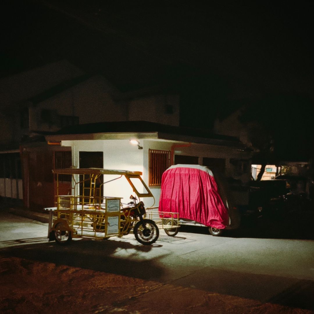A motorbike with yellow utility racks sits in a pool of light next to another small vehicle under a red cover. White houses are visible behind them.