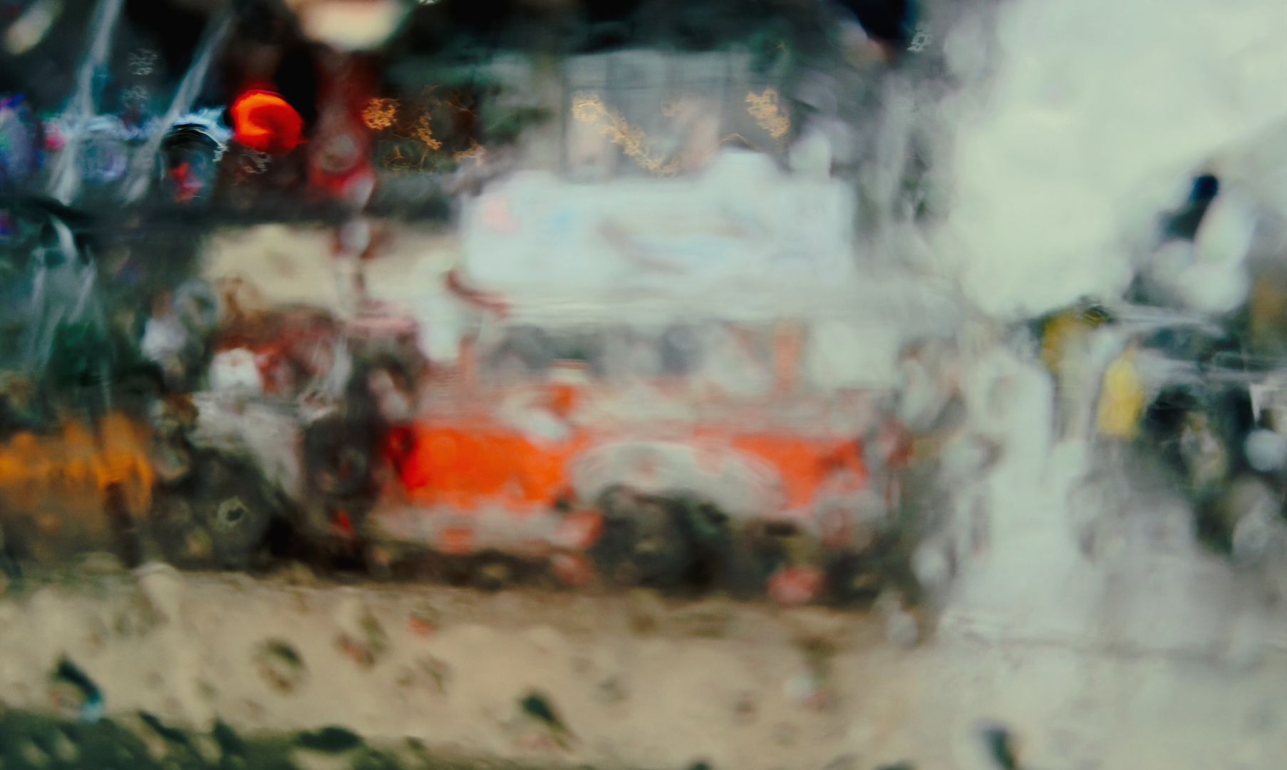 A blurred view through a wet window. Colours run together in this wet street scene. A red jeepney appears in the scene.