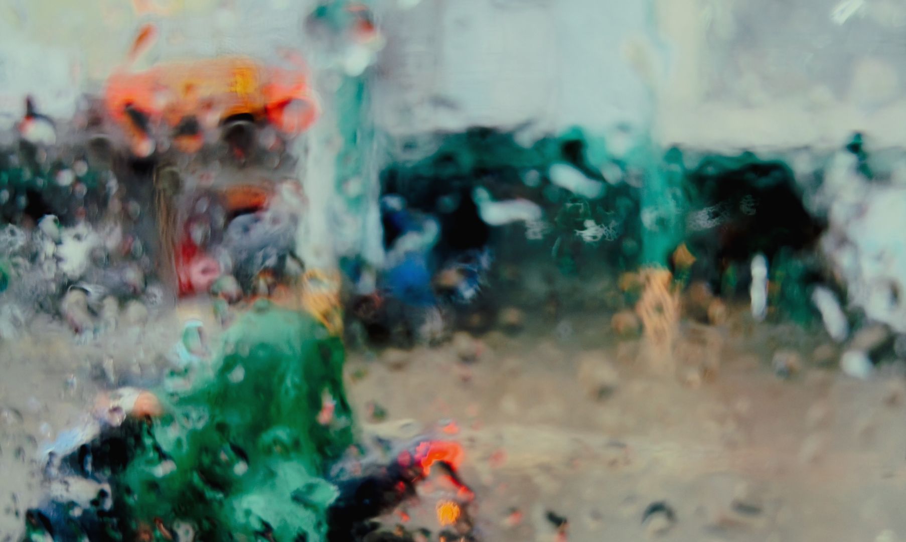 A blurred view through a wet window. Colours run together in this wet street scene. A rider appears in the foreground on a bike wearing a green poncho.