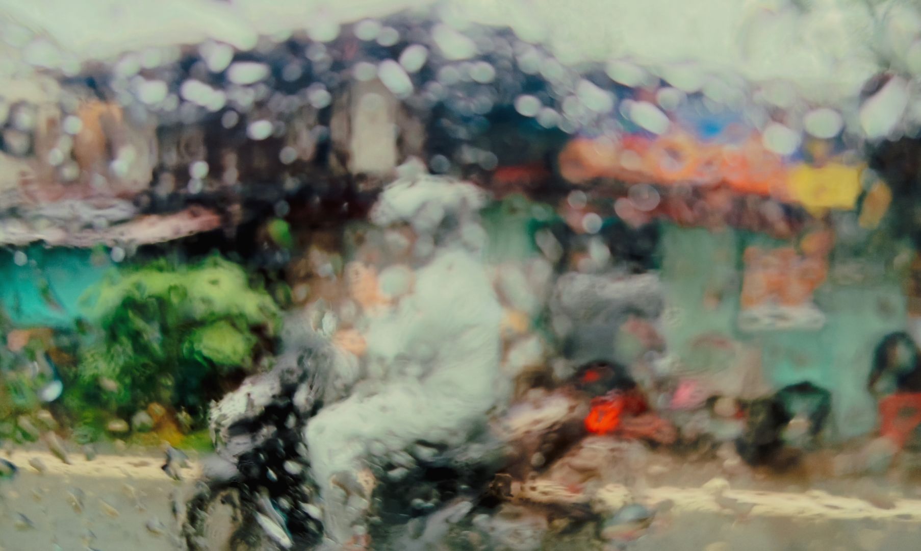 A blurred view through a wet window. Colours run together in this wet street scene. A motorbike rider passes shops on the footpath.