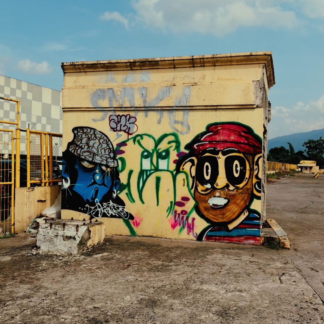 Graffiti mural on a small light yellow building wall, featuring a blue profile face in a patterned cap on the left, green abstract shapes in the center, and a cartoon face with large eyes and a red hat on the right; concrete lot in front with a low block, yellow fence and industrial building to the left, and distant mountain under a partly cloudy sky.