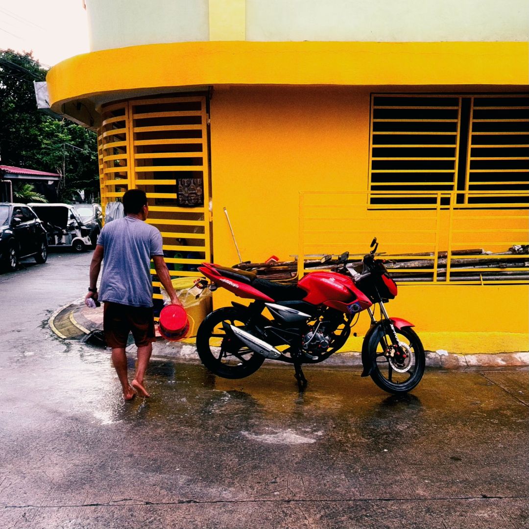 An individual dumps a red bucket in the street next to a red motorbike that is parked in front of t of a bright yellow building.