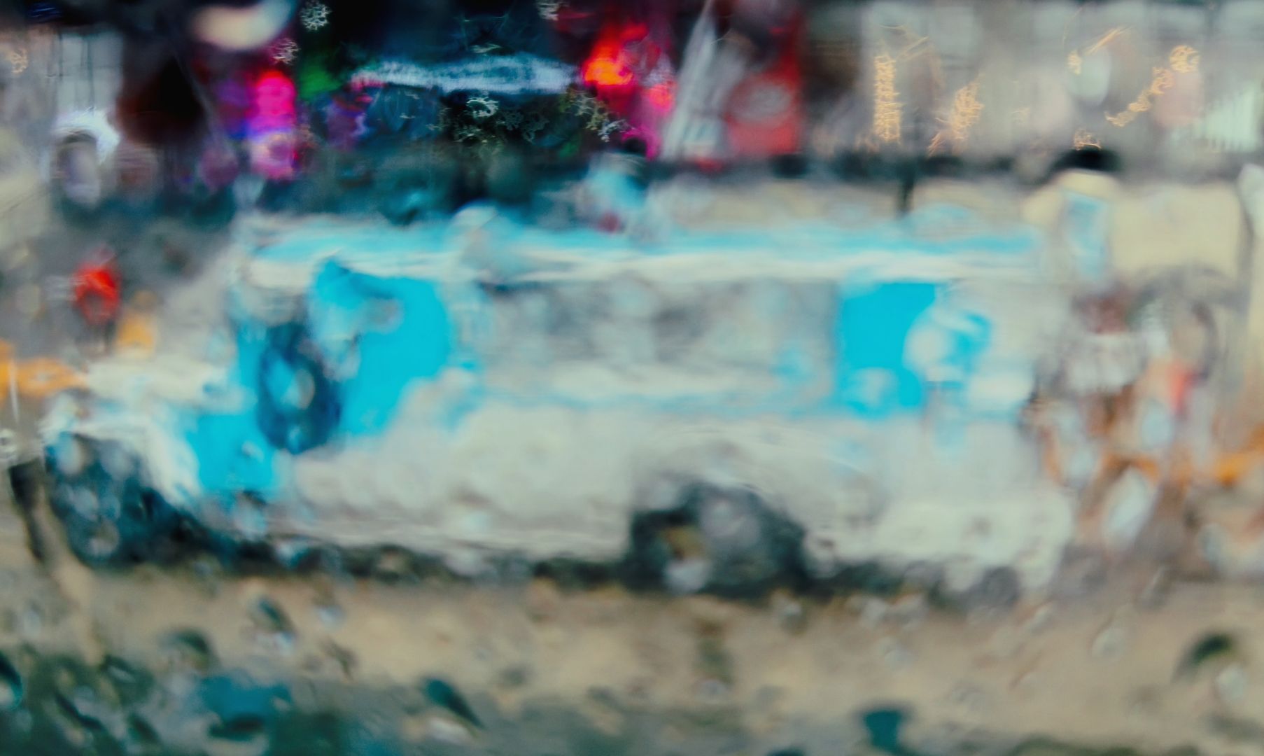 A blurred view through a wet window. Colours run together in this wet street scene. A blue jeepney appears in the scene.