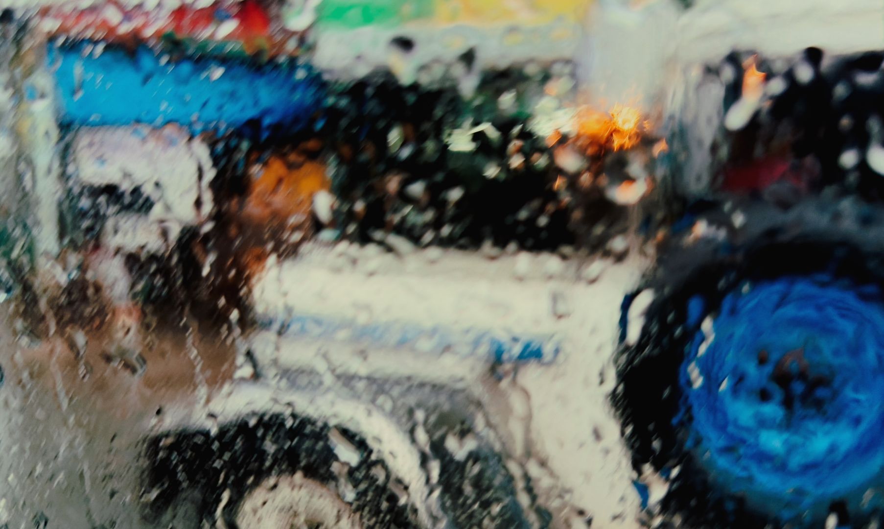 A blurred view through a wet window. Colours run together in this wet street scene. The front of a jeepney is visible, silver with a blue spare tire.