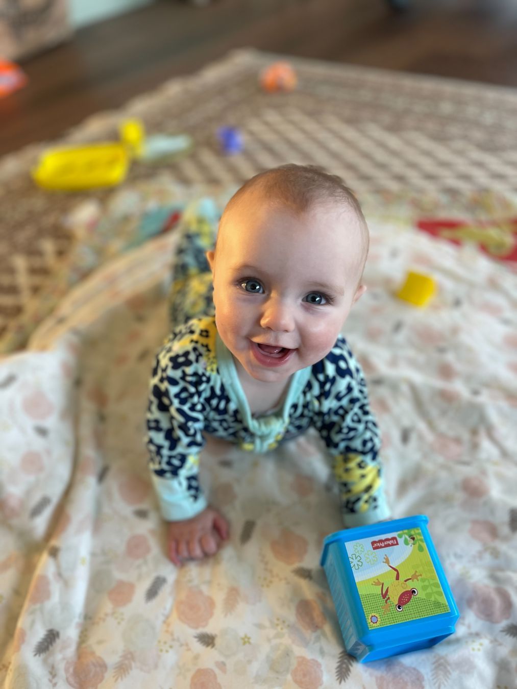 Babby, laying her in belly on a playmat. In her left hand is some weird plastic cube thing