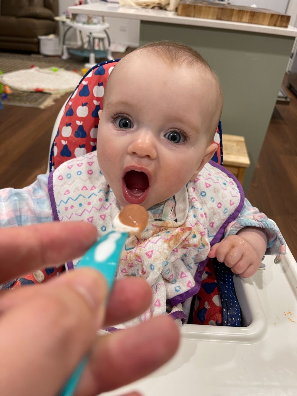 Babby sitting in a high chair. Her mouth is wide open and a spoon of baby goop is about to go in there.
