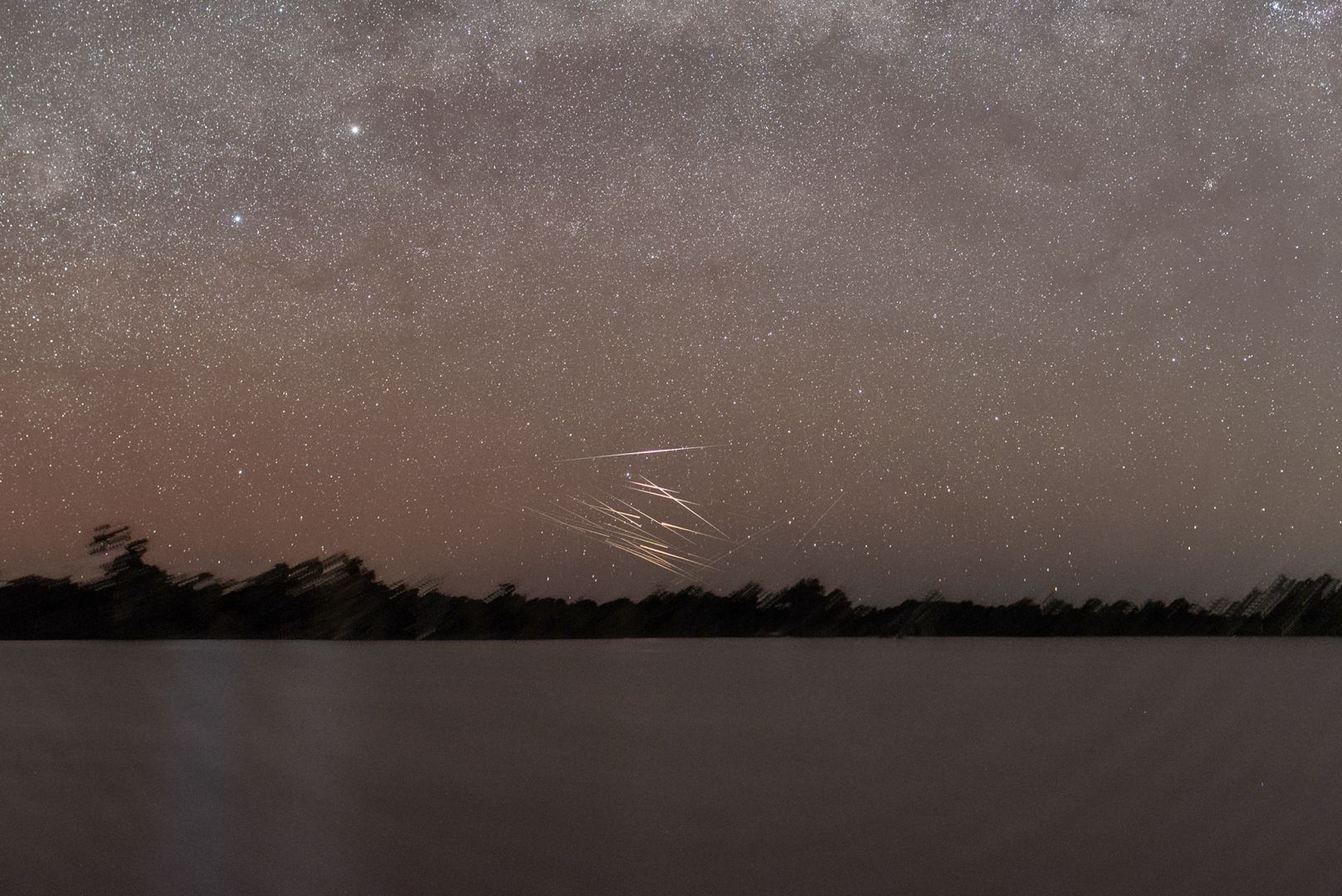 Nightscape view across a lake. Two bright stars (alpha and beta Centauri) are visible towards the left and the band of the setting Milky Way stretches from left to right.

Just above the tree-line across the lake, a large amount of bright streaks are visible. There several sets of parallel streaks and each parallel set is at an angle to the other sets.
