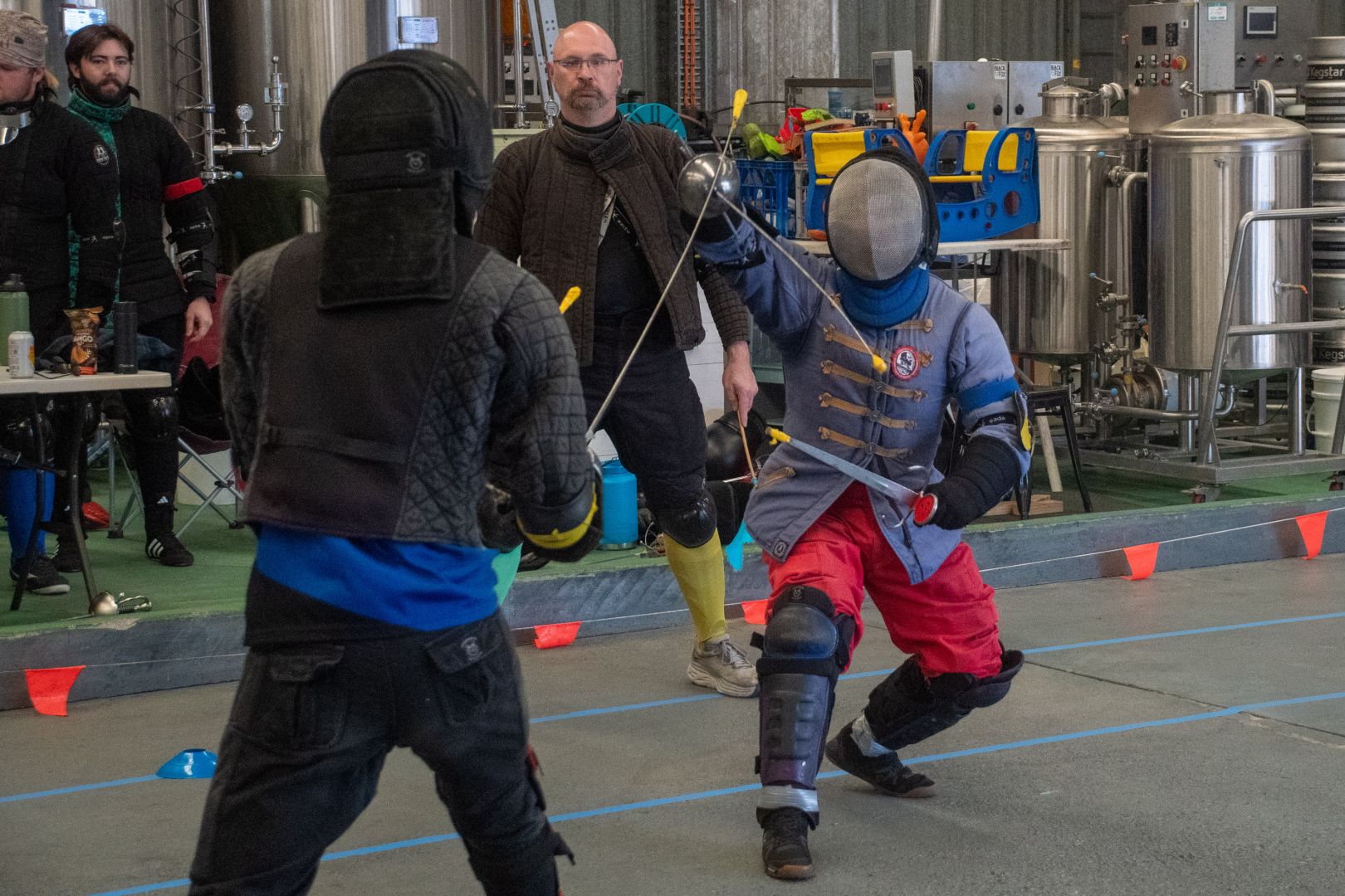 Two fencers in HEMA jackets with rapier & dagger. The one on the right parries a cut to his head with his rapier. In the background is a flag judge, and behind that is brewery equipment.