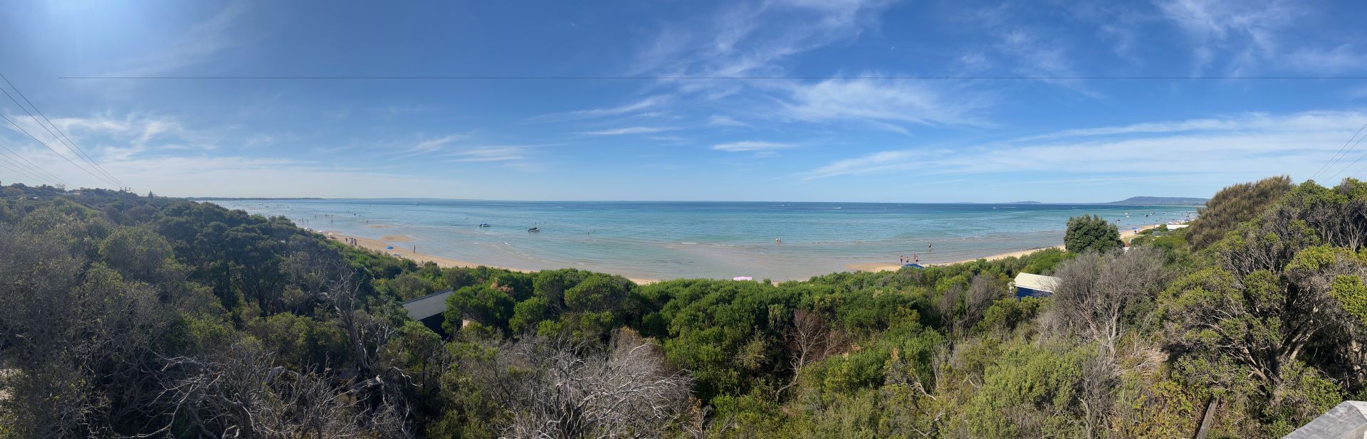 A panoramic view of Rye's front beach in summer. The sky is mostly clear. The tops of trees lining the beach are in the foreground. There are a number of small motorboats in the water as well as people enjoying the a splash.