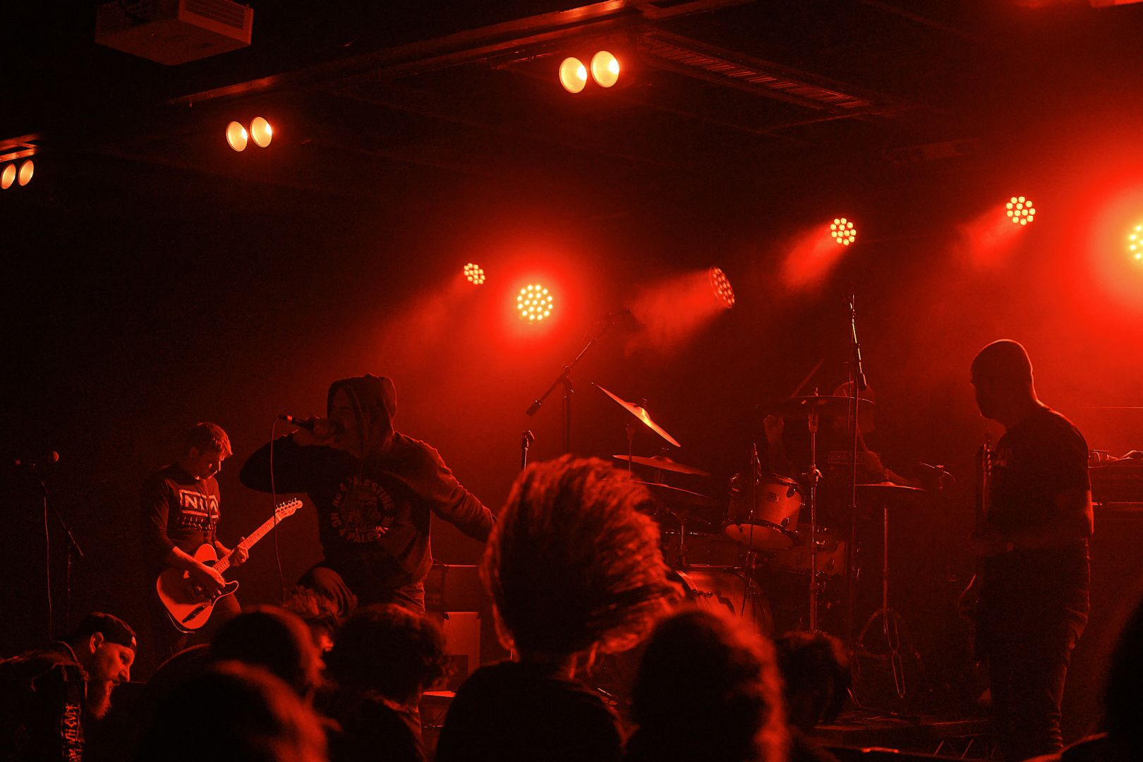 A few crowd members in the foreground headbang while slowcut throwdown on stage. Intense red lighting bathes the whole scene.