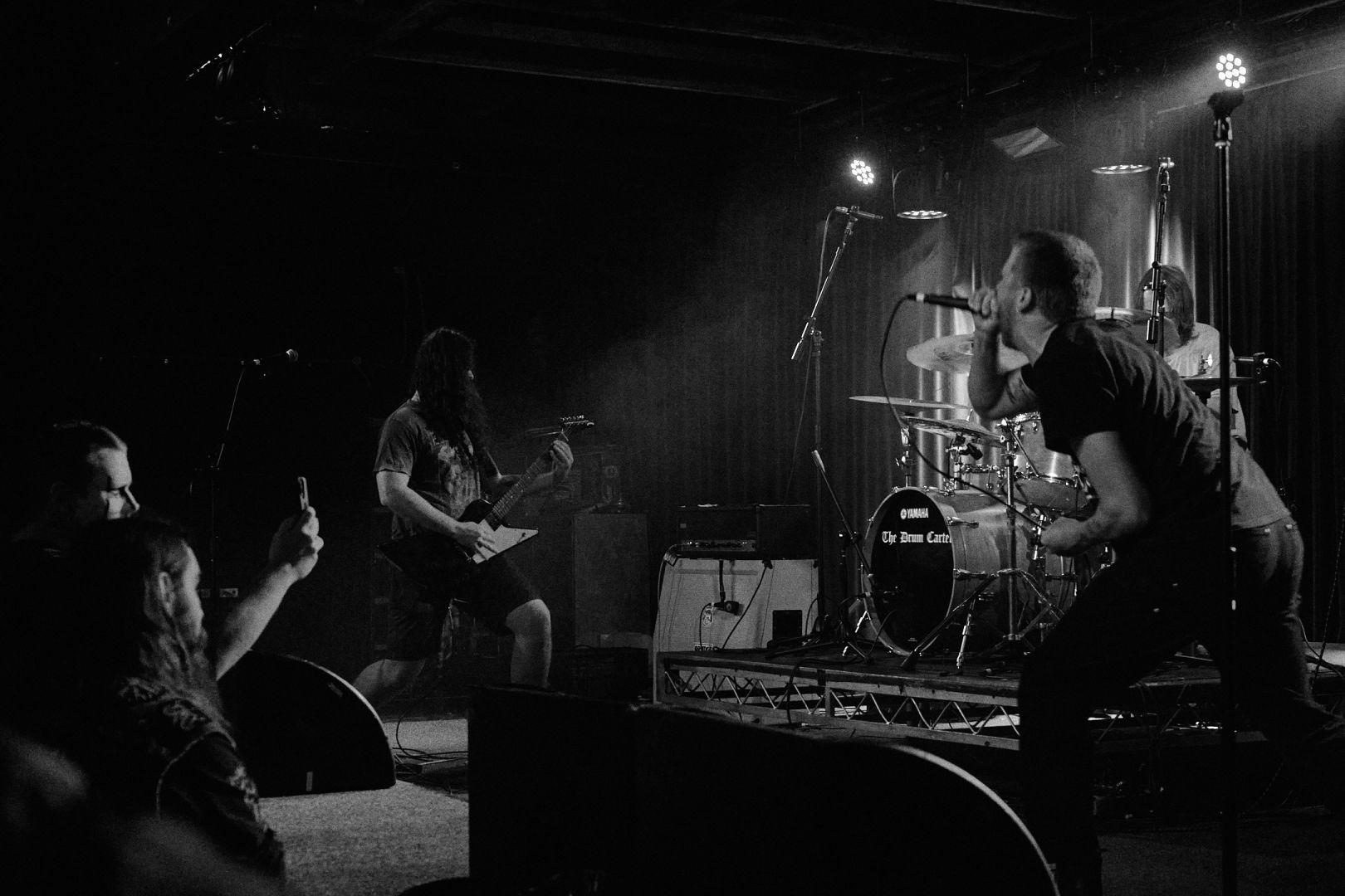 A black and white image of Cerebral Erosion performing on stage. Image right is the vocalist with the drummer behind him, image left is the guitarist and a crowd member taking a snapshot.