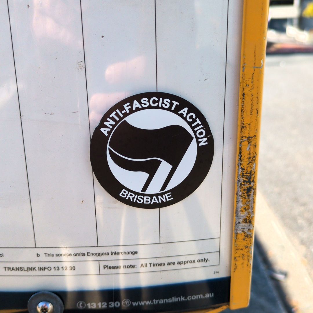 A close up photo of a circle sticker on a bus stop. Text around the outside reads "antifascist action Brisbane", and inside the circle two black flags are waving.