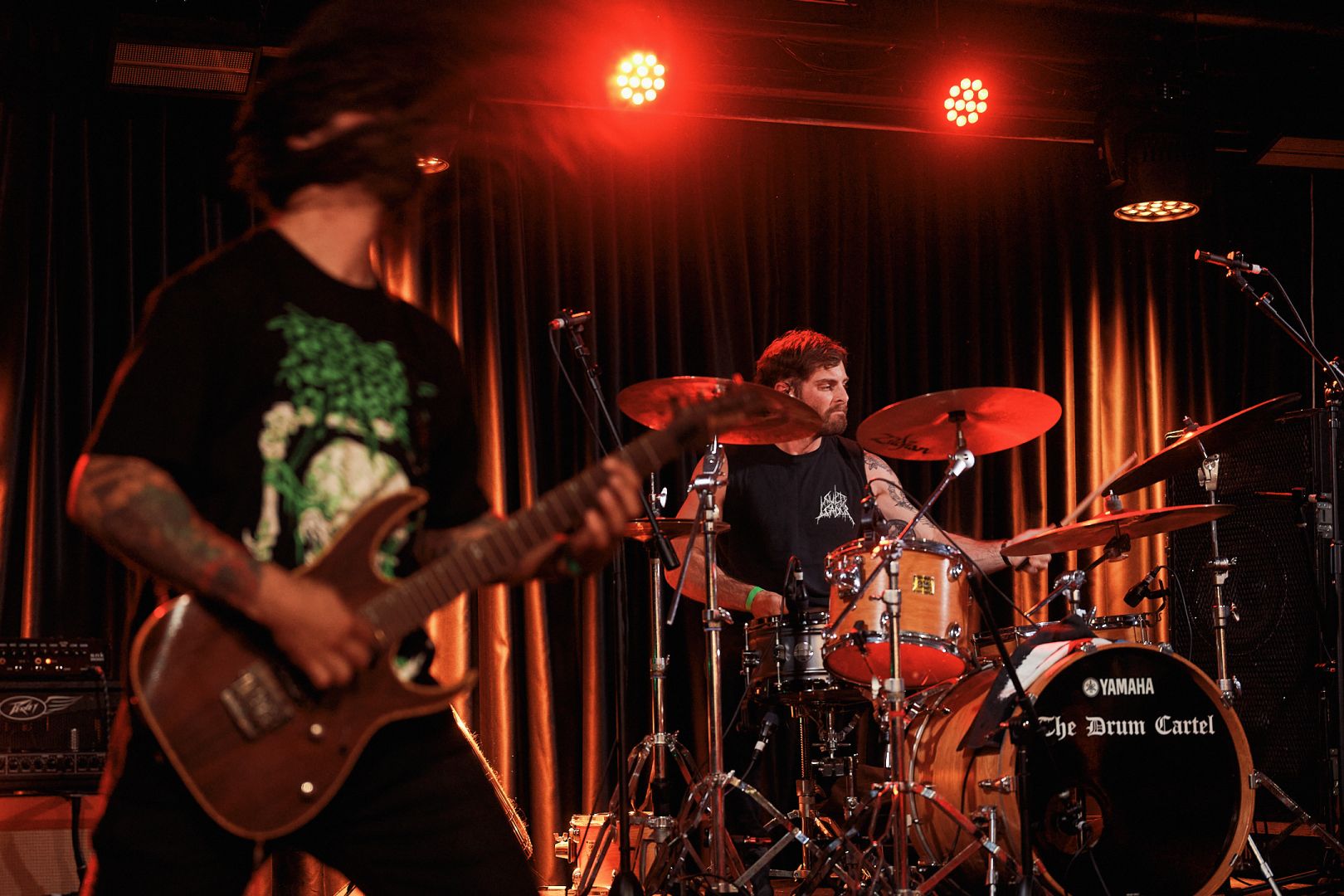 Resin Tomb's guitarist throwing some sick windmills in the foreground, with the drummer behind him bathed in red stage lights.