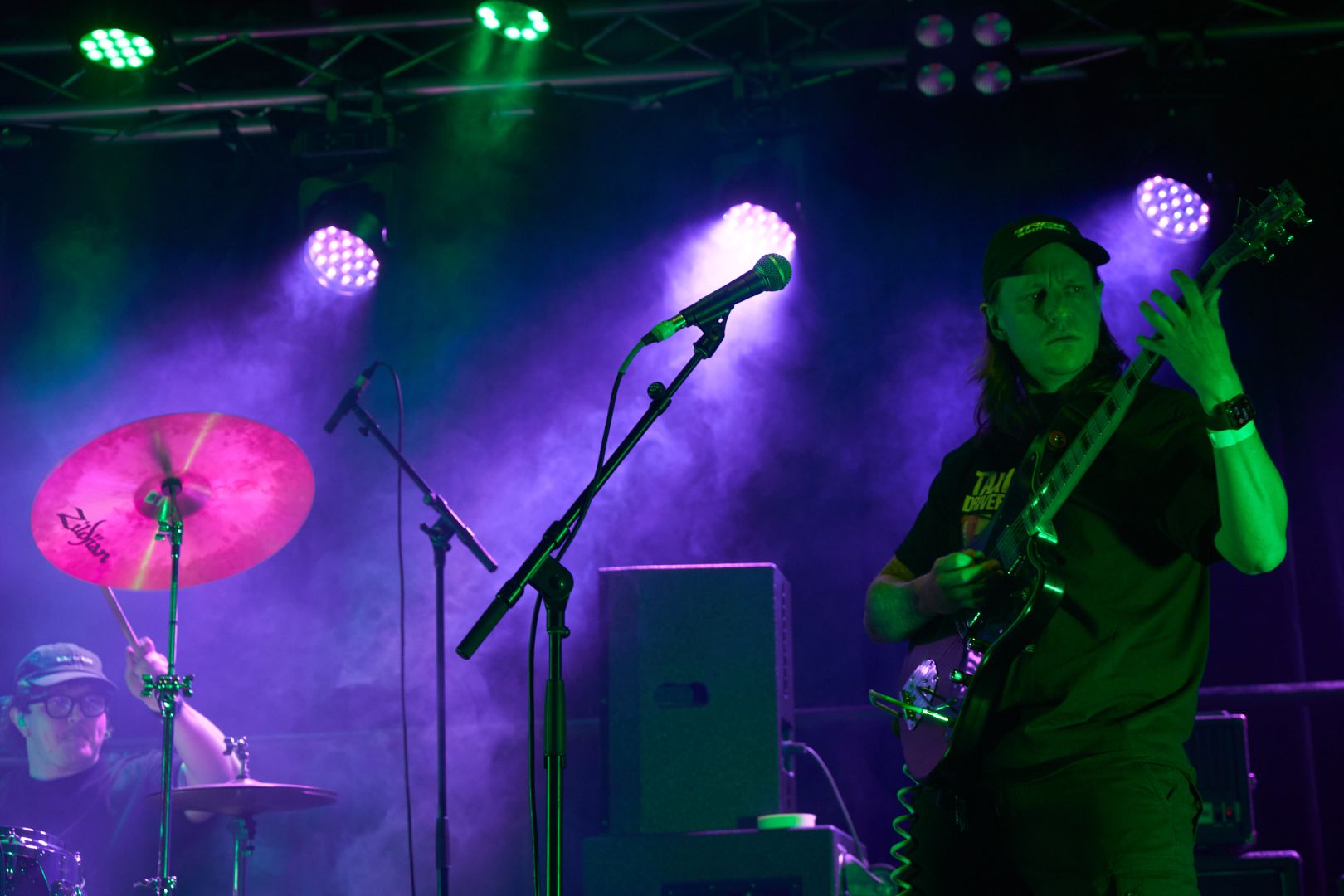 The guitarist in the right-side foreground, washed in green light, attends to the task at hand. The drummer is behind shrouded in what could be fog or mystique.