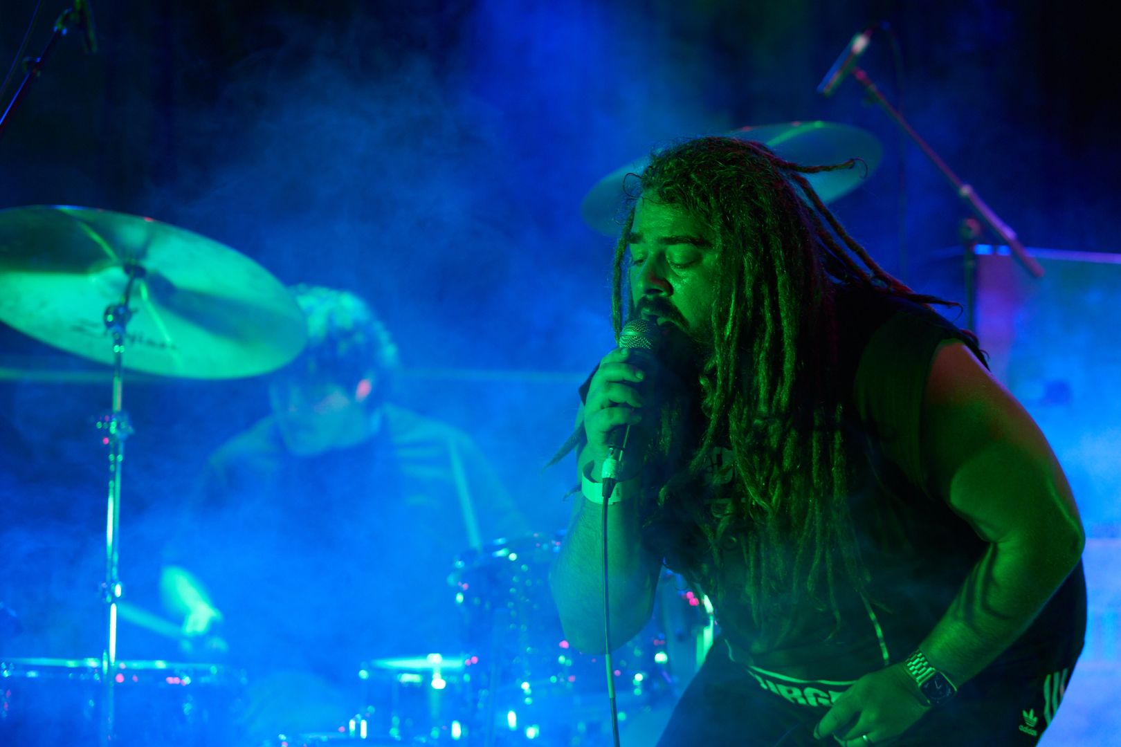 Vocalist is crouched in front of the drummer, with a green and blue wash of light through the fog.