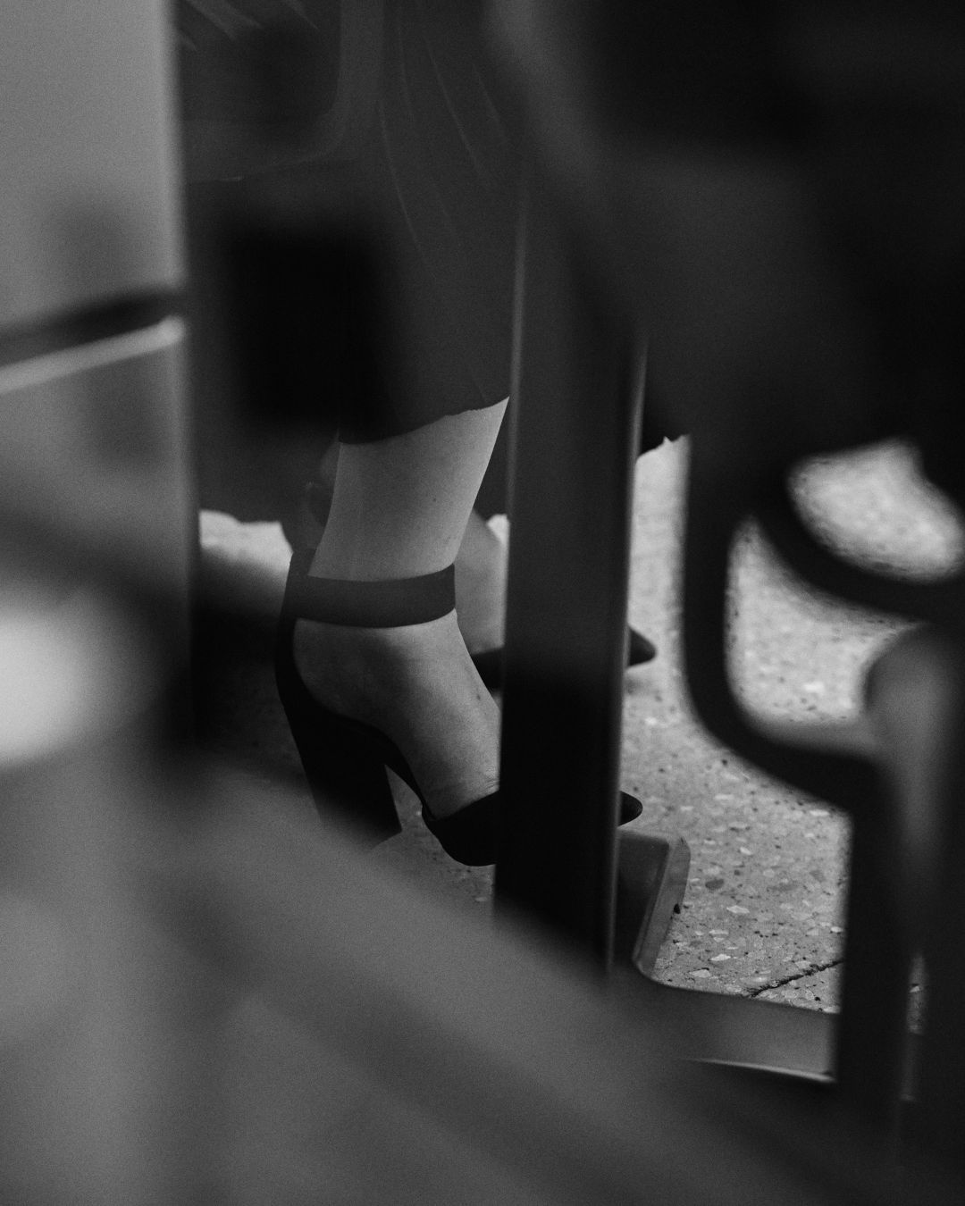 A black and white detail shot of a woman's feet. She is wearing strapped heels and sitting at an outdoor cafe chair.