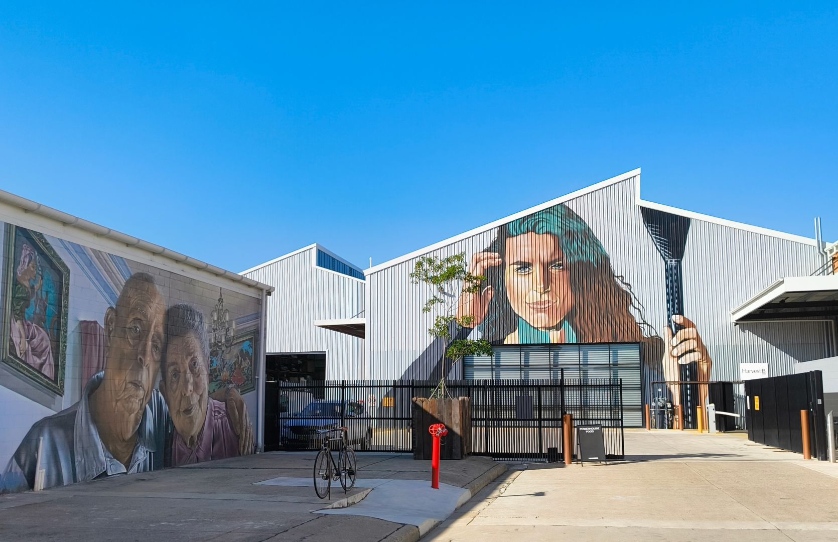 The front of the Harvest B Foundry building under a clear blue winter sky. A huge mural of a woman holding a weaponlike doodad is painted on the building. The building to the left has a different mural of an elderly couple.