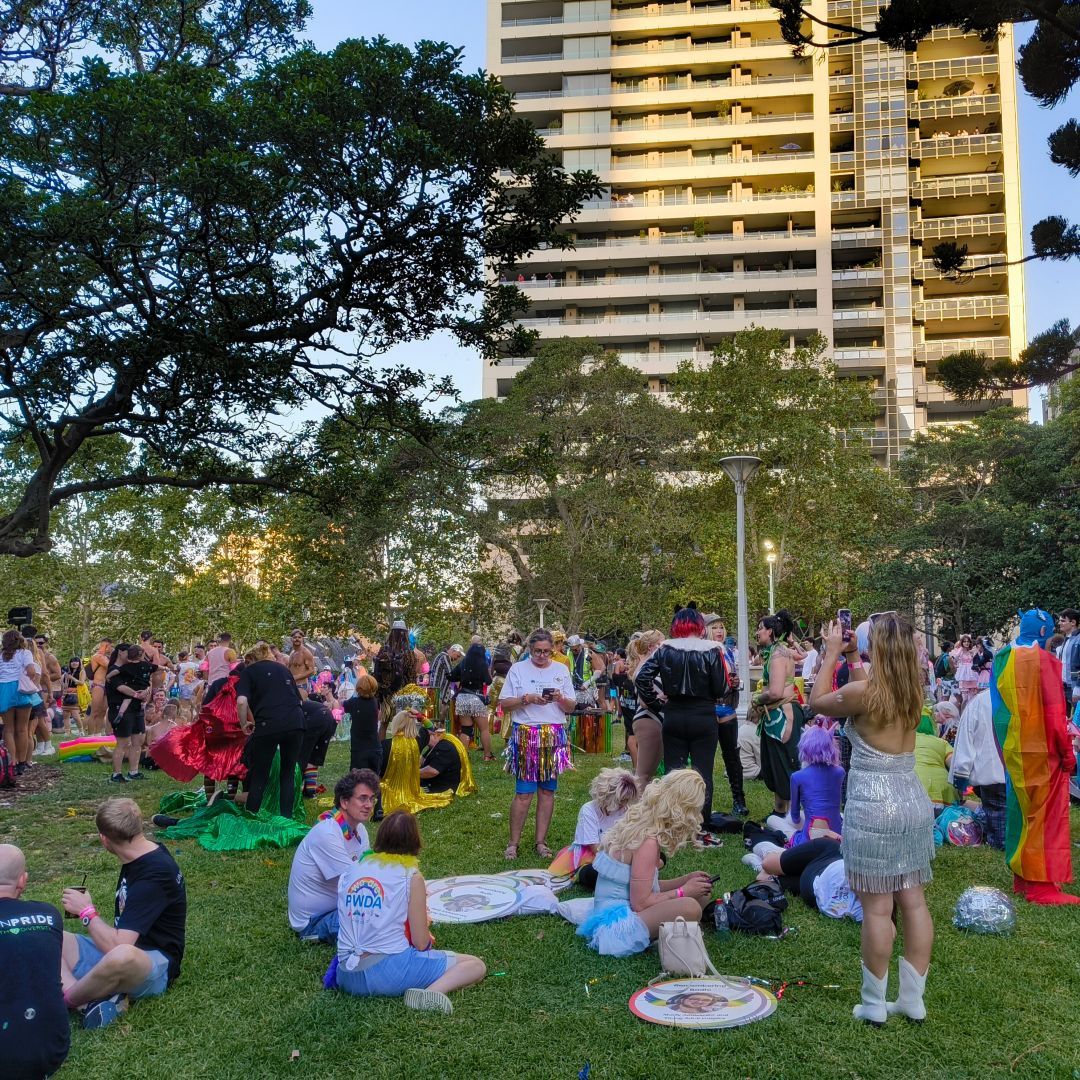A wide shot of Hyde Park before the Sydney Mardi Gras parade. Lots of colourfully dressed people are milling around in groups.