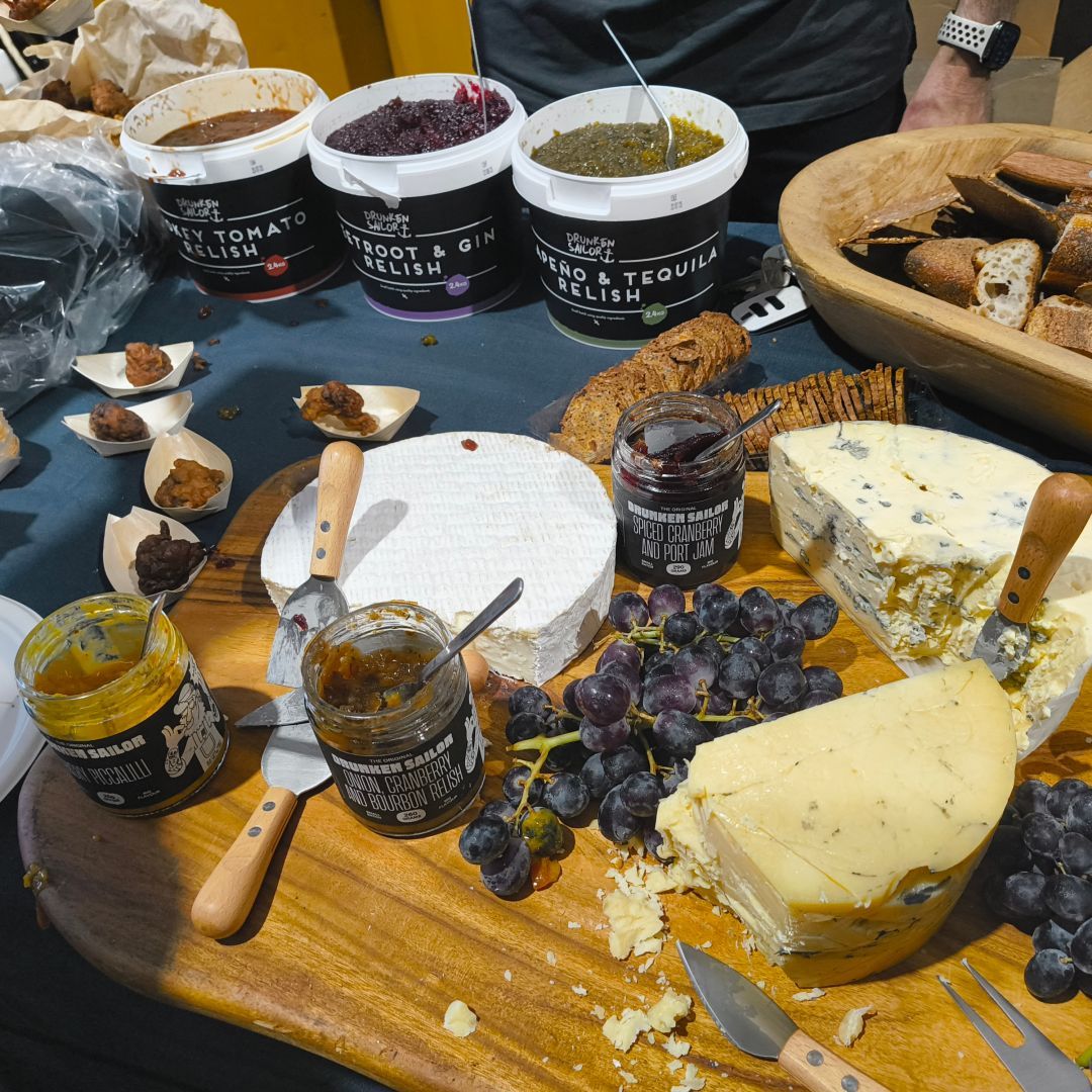 A spread of relishes and jams made by Drunken Sailor Canning Co with fresh grapes, multiple big hunks of cheese, fried zucchini, bread and crackers.