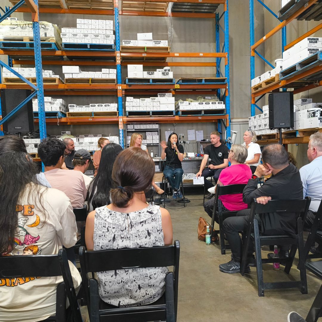 A group of people listening to a panel discussion in the Drunken Sailor Canning Co warehouse. Everyone is sitting on folding chairs and the shelves are stacked with jars of pickles and jams.