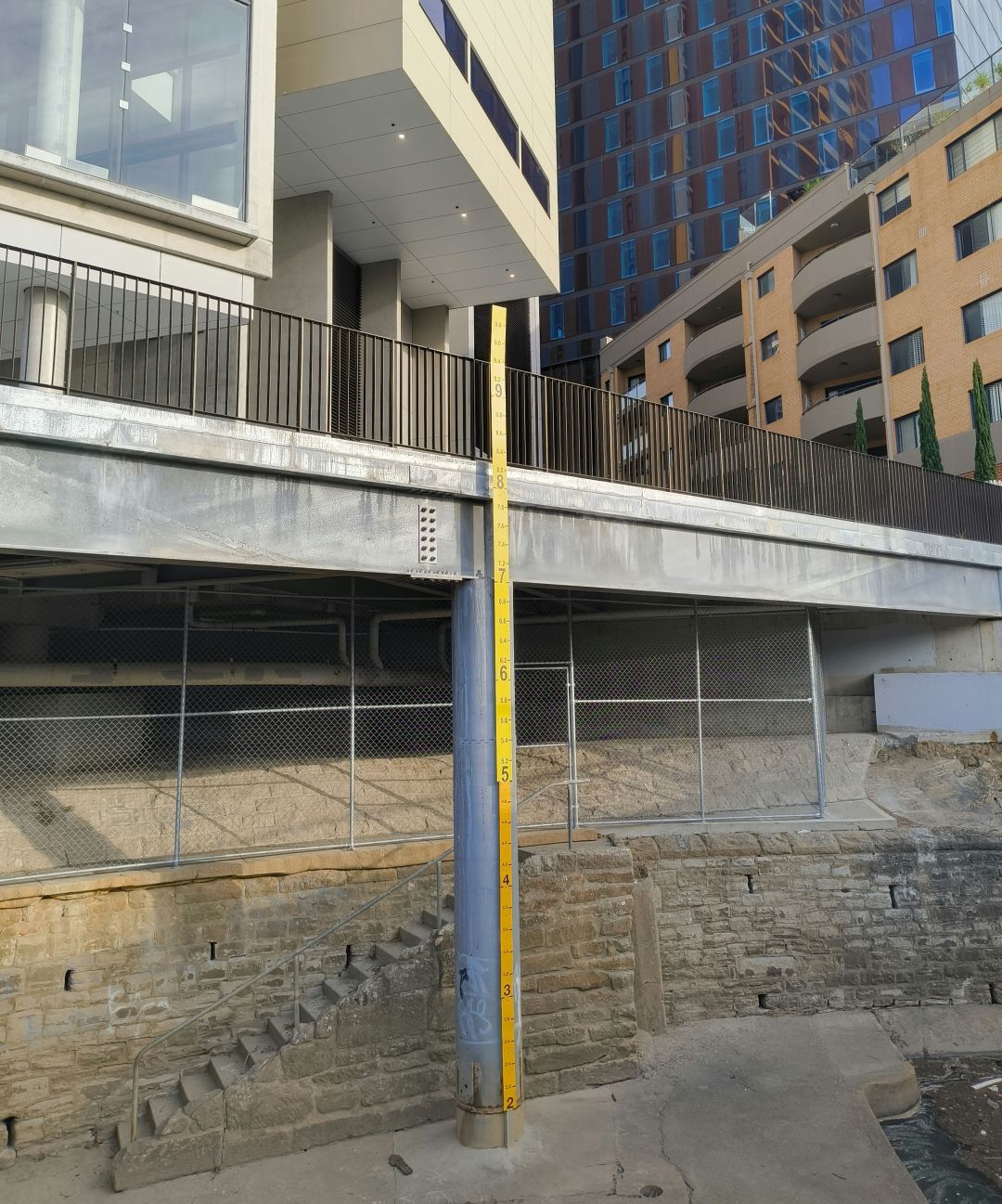 A photo of a flood marker under a high rise building next to Parramatta River. The bottom half of the marker ending at 5m is a different colour to the top half ending just above 9.8m.