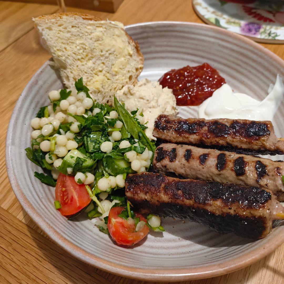 Homemade dinner with grilled lamb koftas, pearl couscous salad, various dips and a slice of sourdough bread.