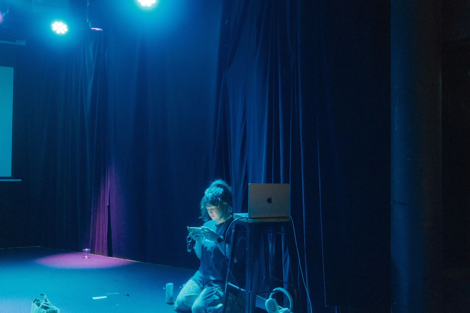 comedian demi lardner sits under a blue light, checking her phone on stage, immediately after announcing her show is complete