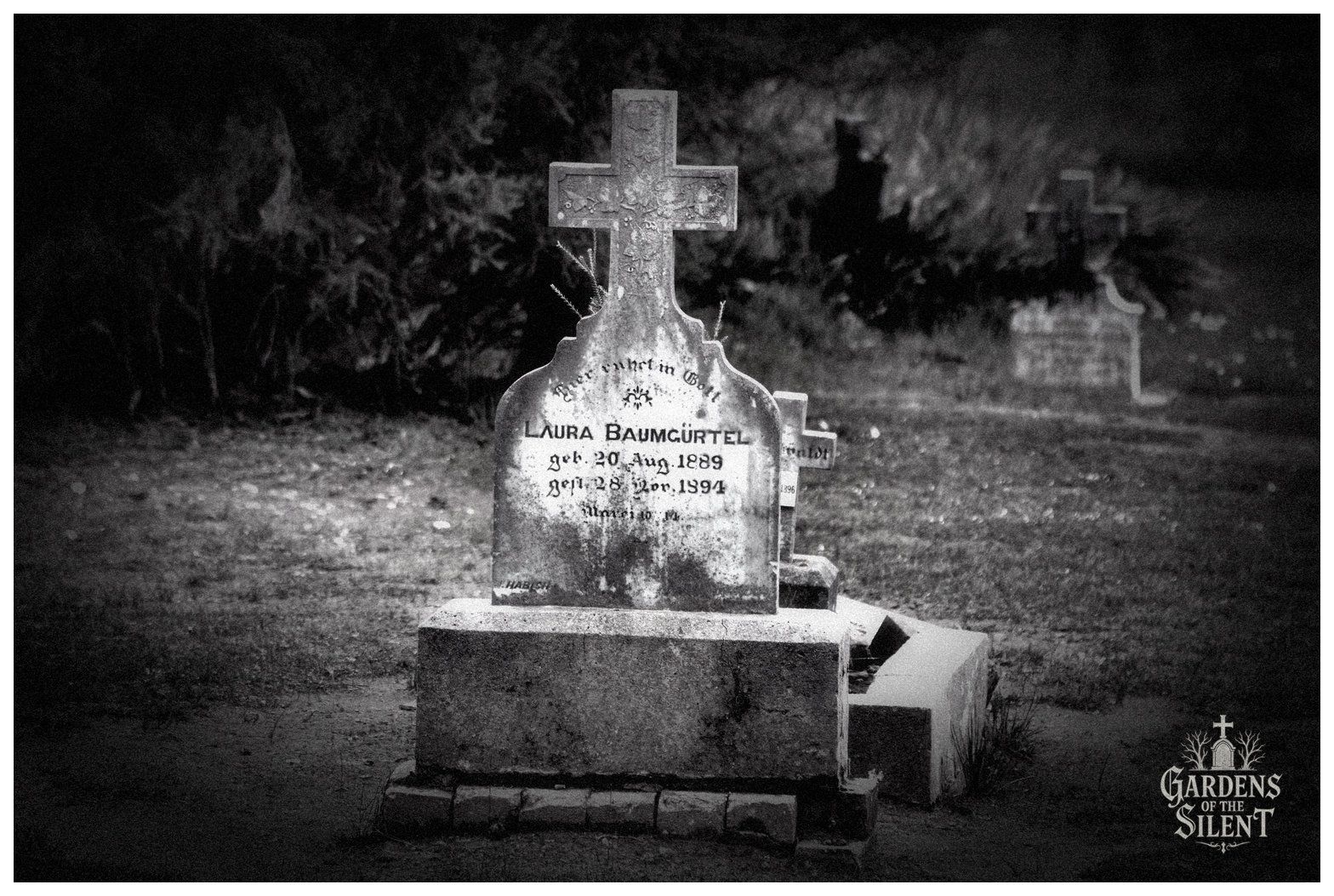 Black and white photograph of an ornate, weathered stone headstone commemorating Laura Baumgürtel, who lived from 1889 to 1894. The headstone features a carved stone cross atop a curved shoulder tablet resting on a solid rectangular pedestal. The weathered surface shows heavy dark patina and German script. In the soft focus background, dark foliage and another distant cross headstone stand out against the lawn. The watermark Gardens of the Silent appears in the lower right corner.
