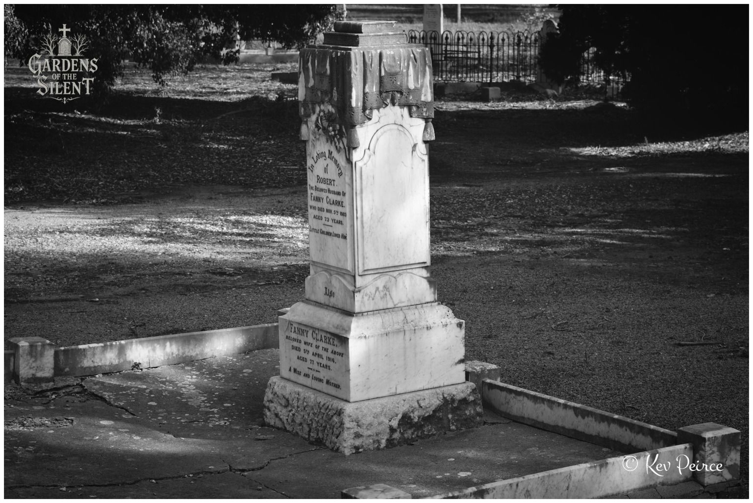 A black and white, eye level shot of an ornate marble pillar headstone for Robert Clarke and Fanny Clarke. The monument features meticulously carved drapery and tassel details around its upper cap. Strong sunlight casts distinct shadows across the stone surfaces, concrete grave border, and the open cemetery lawn in the background.