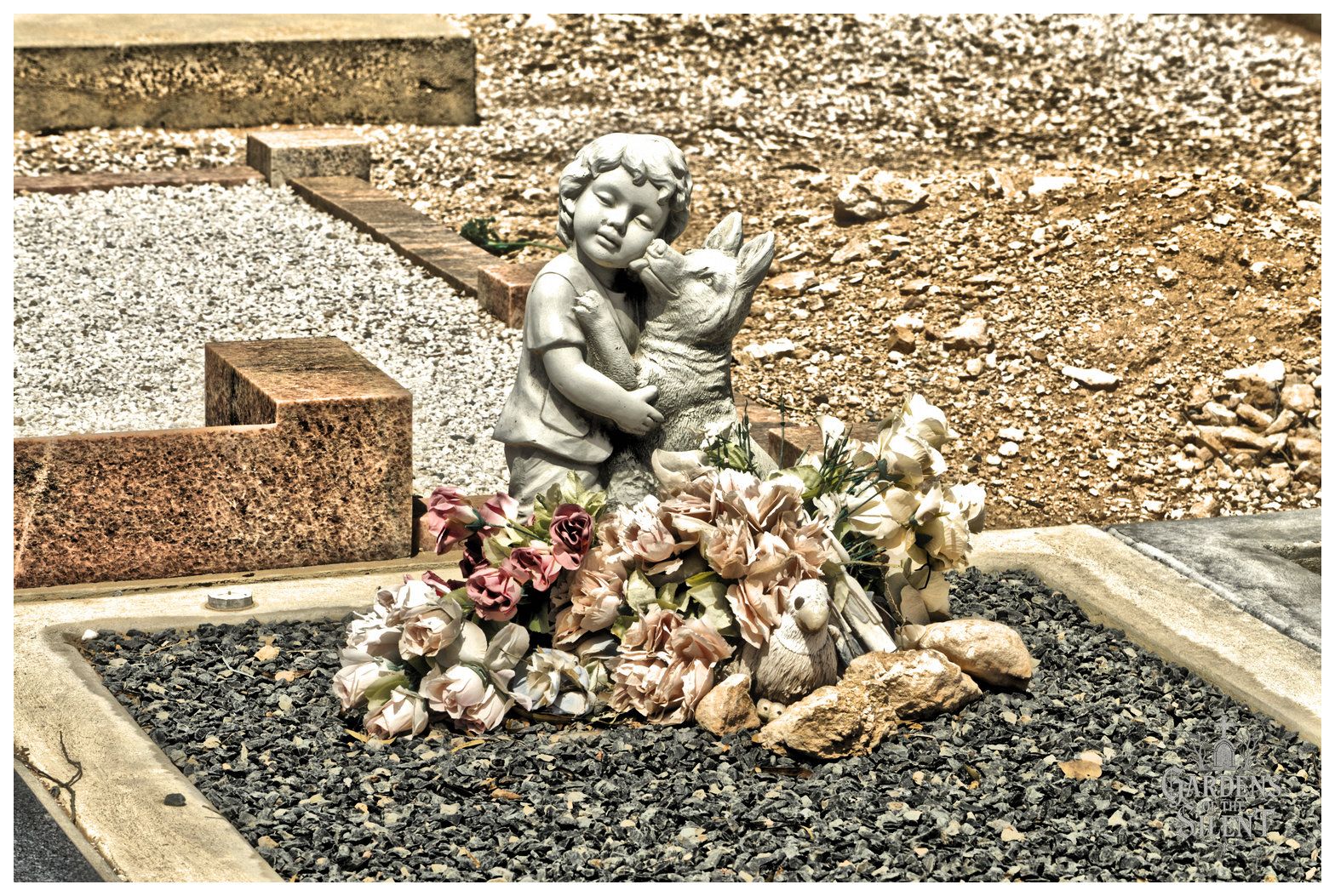 A colour, high contrast close up photograph of a grave plot covered in dark grey gravel. In the centre sits a carved stone statue of a young child lovingly hugging a dog that looks up towards the child's cheek. At the base of the statue rests an arrangement of faded artificial flowers in pale pink, cream, and rose tones, accompanied by a small sculpted bird and rough garden stones. Bright sunlight casts crisp shadows across the pale gravel surround and marble kerbing.