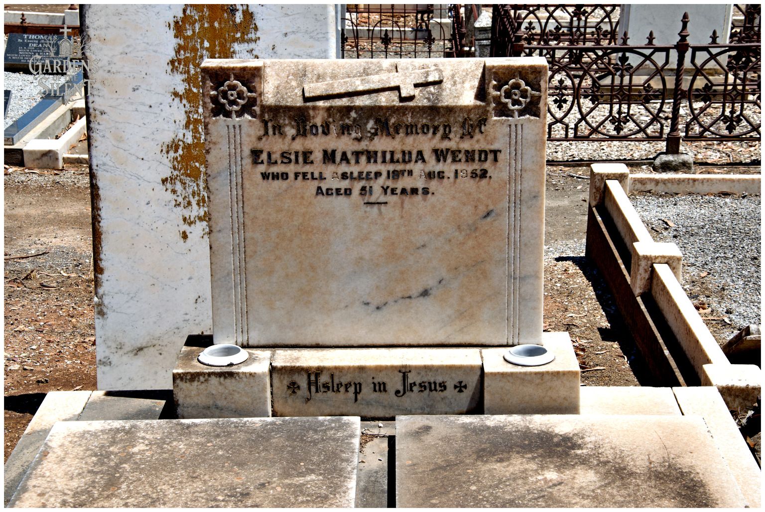 A close up colour photograph of a rectangular marble headstone with a cross carved horizontally across the top section. The inscription is dedicated to Elsie Mathilda Wendt, who passed away in 1952. Two small circular stone holders sit on either side of the base, which reads 'Asleep in Jesus'. In the background, ornate dark iron railings and an adjacent weathered white monument are visible under bright daylight. A watermark reading 'Gardens of the Silent' is in the top left corner.