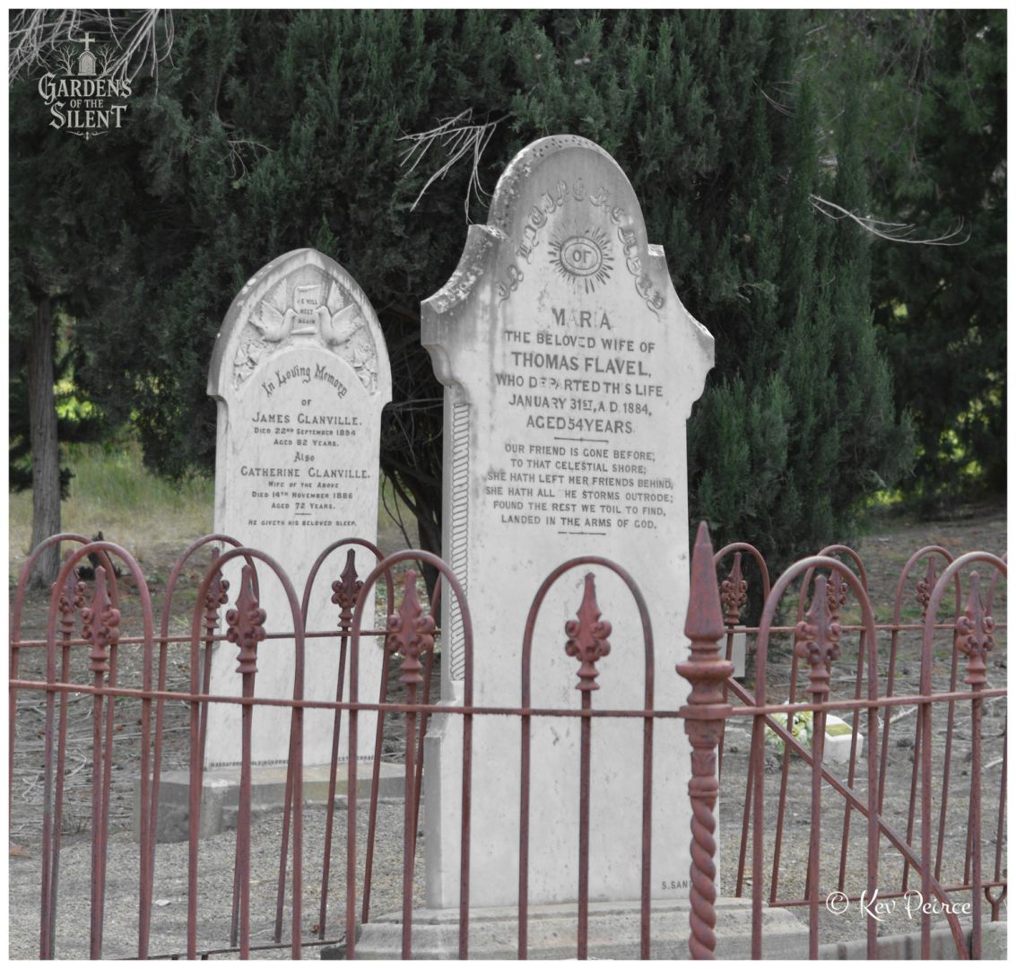 A selective colour photograph framed from behind a weathered, decorative red brown iron fence. Inside the enclosure stand two arched marble headstones, with the prominent front stone commemorating Maria Flavel. Deep green cypress foliage stands out vividly in the background, contrasting with the soft, monochrome stone and gravel path under open daylight.