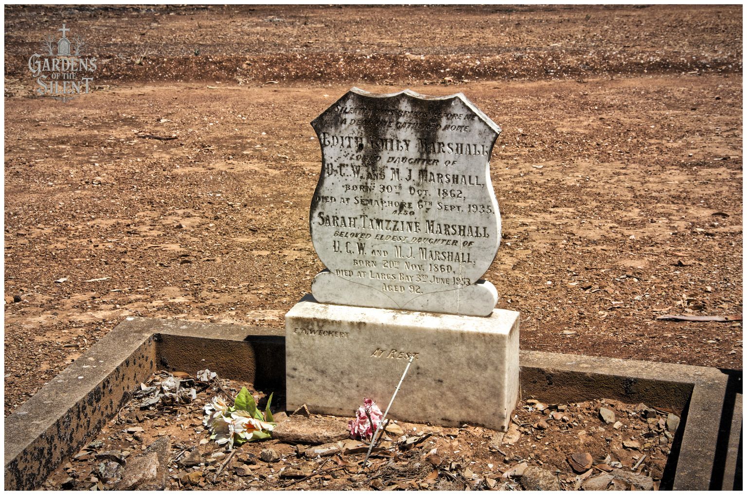 A colour photograph of an upright marble headstone shaped like a shield, set against a background of dry, red brown Australian soil. The headstone commemorates Edith Emily Marshall and Sarah Tamzzine Marshall. A simple concrete border surrounds the dry grave plot, which contains a small bunch of faded artificial flowers and a thin stick leaning against the base. Sunlight casts a soft shadow to the right. A watermark reading 'Gardens of the Silent' is visible in the top left corner.
