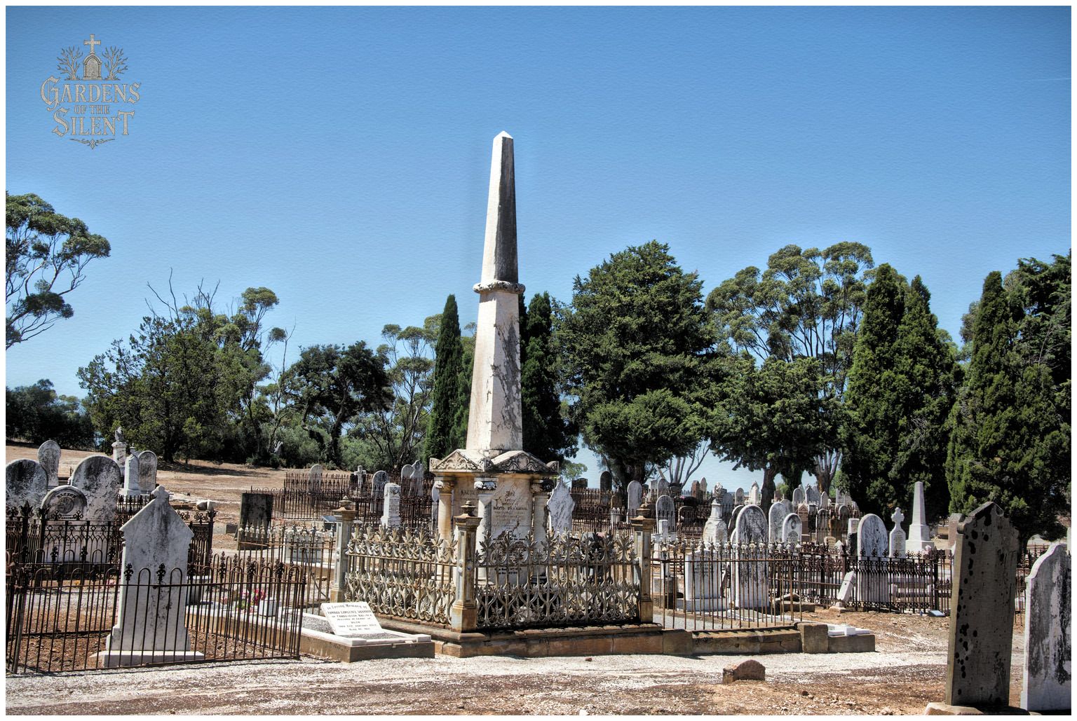 A wide angle colour landscape photograph of a historic cemetery under a clear blue sky. A tall, weathered white stone obelisk stands prominently in the centre, surrounded by a highly decorative cast iron fence. Numerous other white headstones, iron enclosures, and grave plots stretch across the dry ground, bordered by green eucalyptus and cypress trees in the background. A watermark reading 'Gardens of the Silent' is positioned in the top left corner.