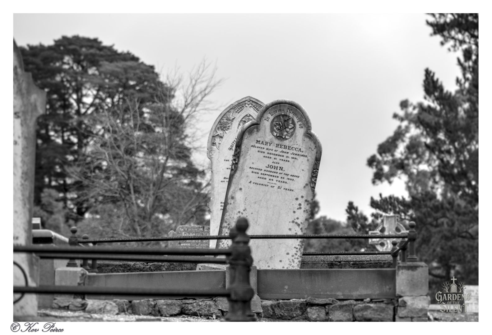 Black-and-white photograph taken by Kev Peirce for Gardens of The Silent, showing two prominent headstones in Norton Summit Cemetery, South Australia. The foreground features the grave of Mary Fenwick (died 23 July 1913, aged 78) and her husband John W. Fenwick (died 8 July 1916, aged 83). The headstones are framed by a wrought iron fence, with trees and other gravestones in the background. The image evokes themes of enduring love, historical memory, and quiet reflection.