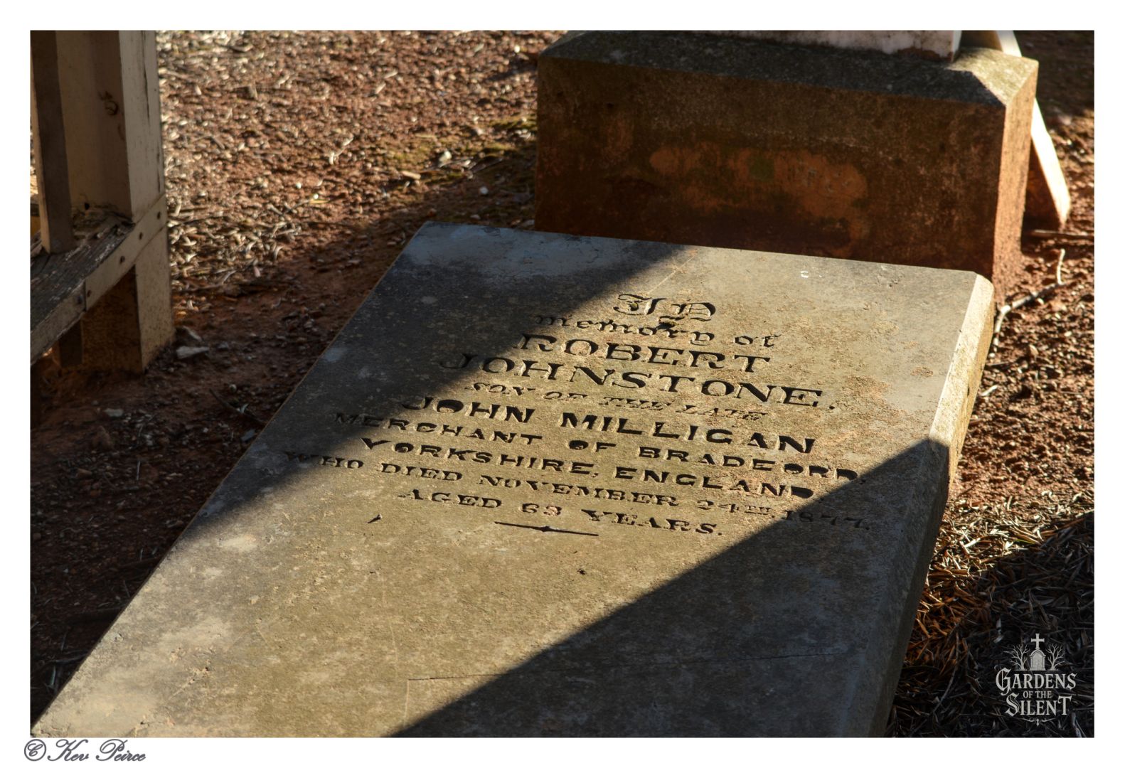 Flat gravestone in Burra Cemetery commemorating Robert Johnstone, son of John Milligan, a merchant from Bradford, Yorkshire. He died on 6 November 1865 at age 63. The stone lies on bare earth, with shadows cast by nearby structures. Inscription is engraved in serif lettering. Watermark reads “Gardens of the Silent.” Photo by Kev Peirce.