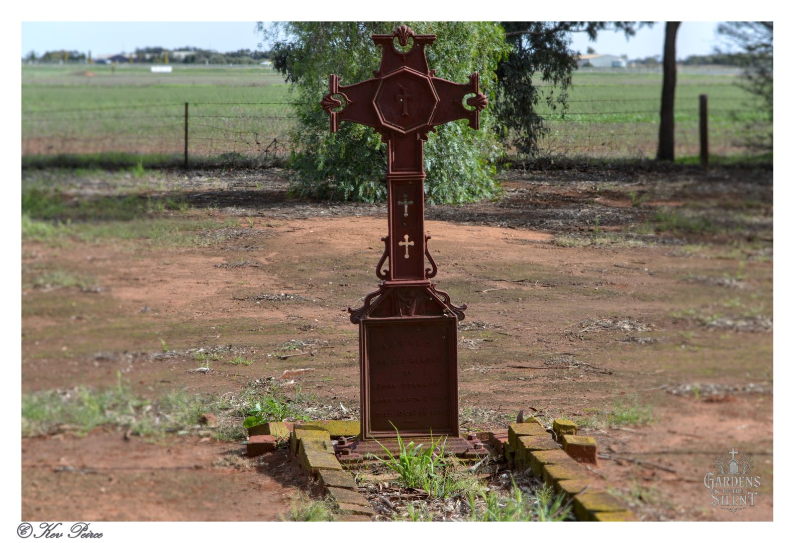 A solitary grave marker stands in a rural field, framed by sparse grass and distant farmland. The marker is a tall, ornate iron cross with intricate detailing and religious symbols, including a central figure of Christ and smaller crosses.  It rises from a brick-bordered grave, weathered and reverent. Behind it, a wire fence separates the sacred from the pastoral, while trees and open land stretch into the horizon.  The image is bordered in white, with “Gardens of the Silent” inscribed in the lower right corner and a signature reading “© Kev Peirce” in the lower left.