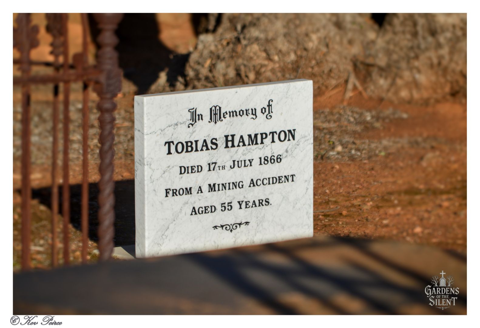 White marble gravestone in Burra Cemetery marking the resting place of Tobias Hampton, who died in a mining accident on 17 July 1866 at age 55. The stone stands on reddish soil with shadows cast across its surface, surrounded by trees. Inscription is clearly legible. Watermark reads “Gardens of the Silent.” Photo by Kev Peirce.