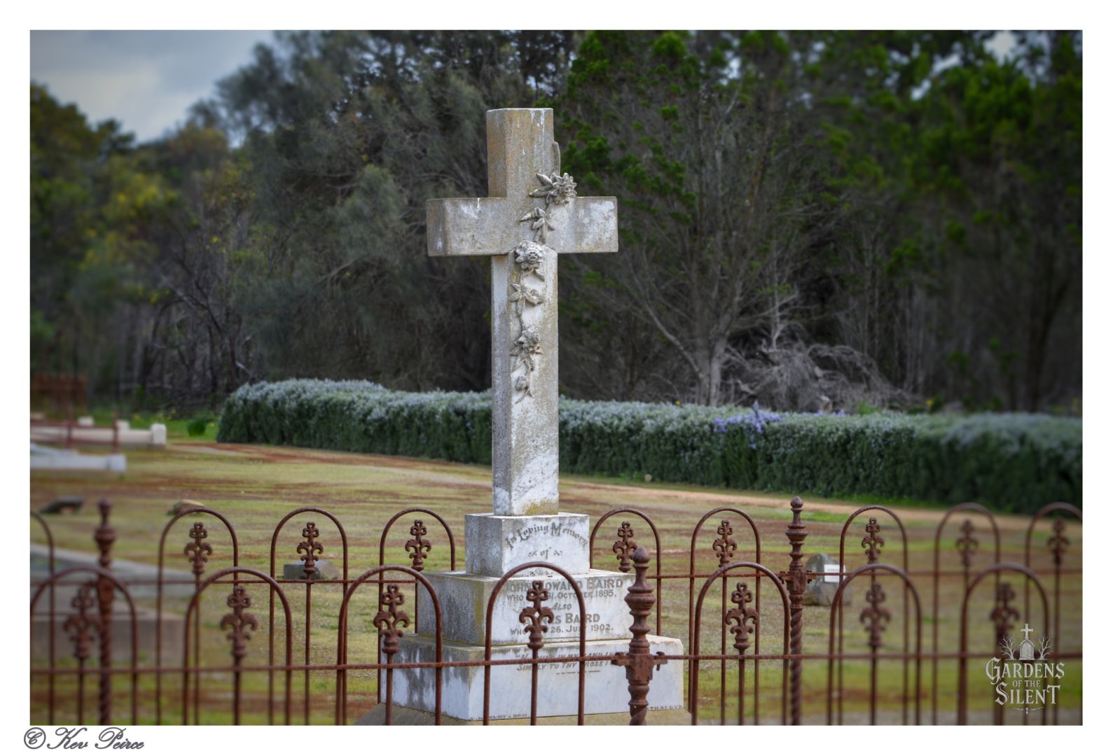 A cemetery scene from Milang Cemetery featuring a prominent stone cross adorned with floral carvings, mounted on a square base with inscriptions.  The monument is enclosed by a decorative iron fence with circular and pointed elements. In the background, additional grave markers and a line of trees suggest a peaceful, well-tended resting place. The sky is soft and overcast.  The image includes the text “Gardens Of The Silent” in the bottom right corner and is signed “Kev Peirce” in the bottom left.